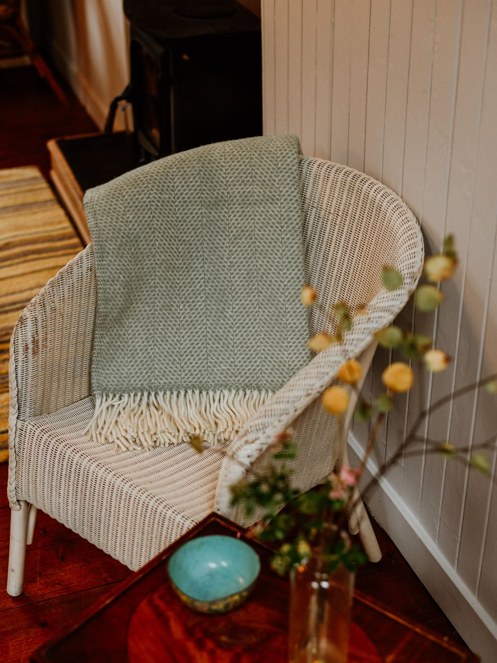 A white wicker chair with a Campbell's of Beauly Stipled Twill Blanket in light green draped over it sits by a wooden table topped with a small blue bowl and vase of flowers in a cozy, warmly lit room.