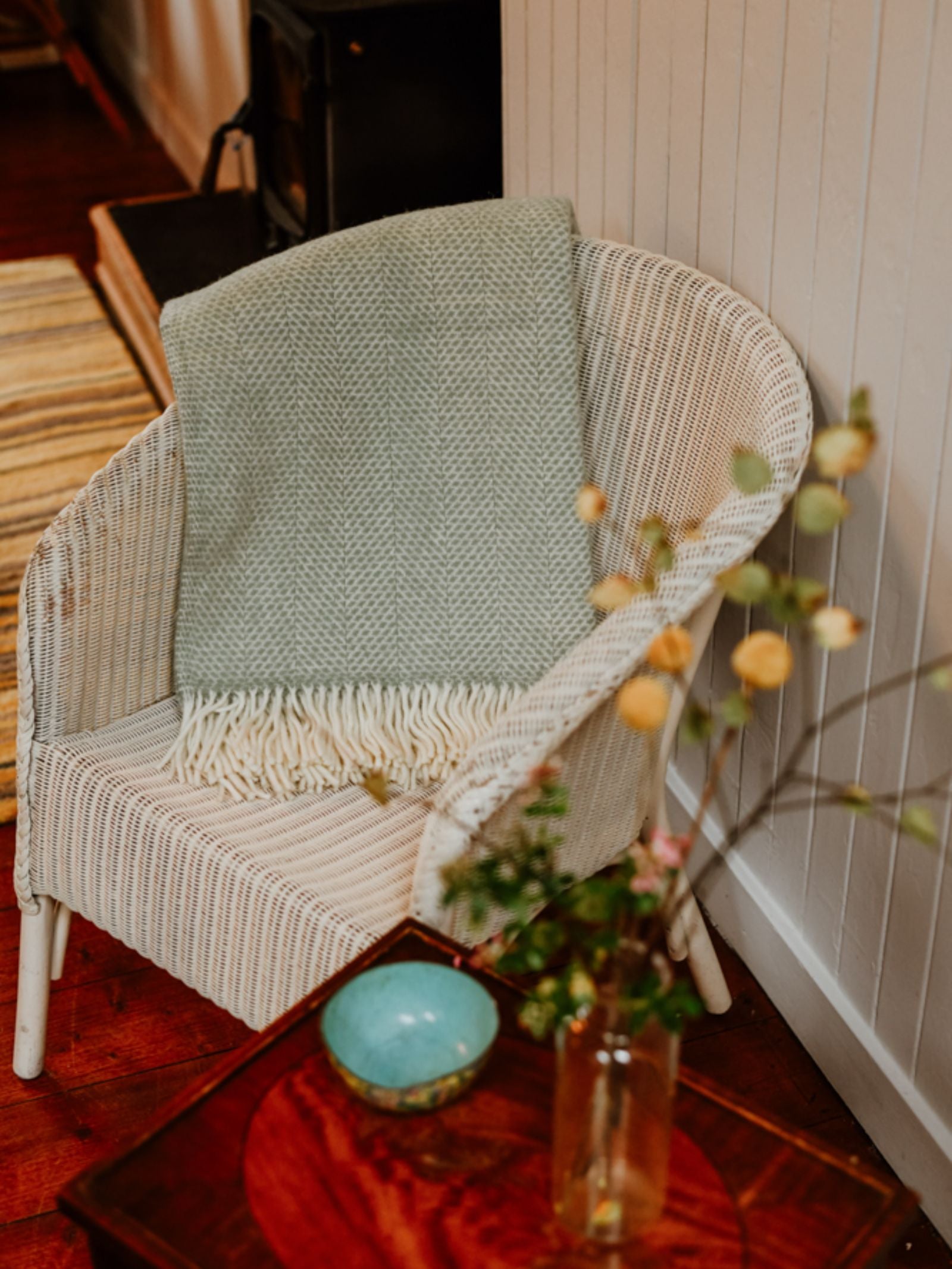 A white wicker chair with a Campbell's of Beauly Stipled Twill Blanket in light green draped over it sits by a wooden table holding a blue bowl and yellow flowers, creating a cozy, warm atmosphere.