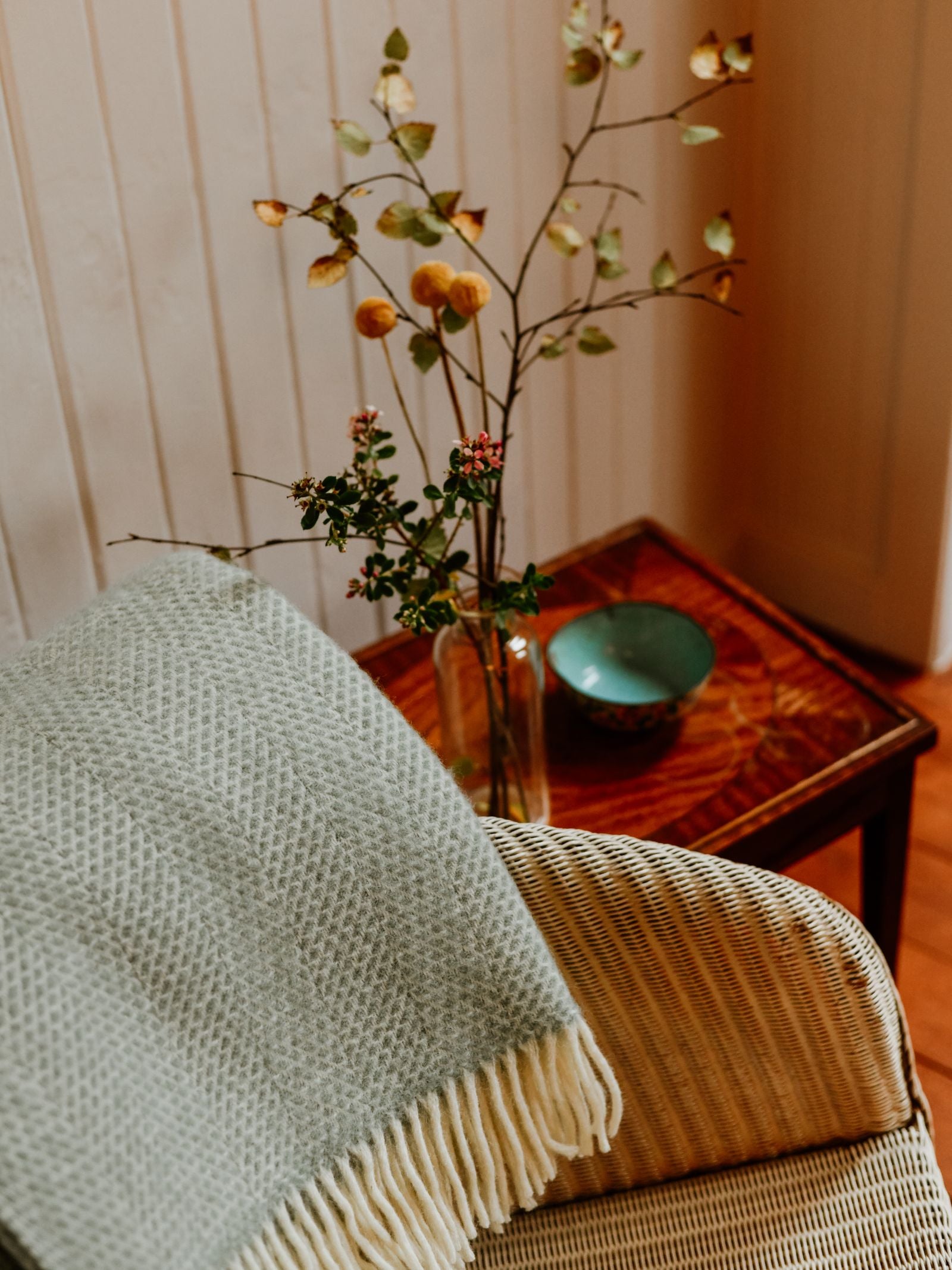 A Stipled Twill Blanket by Campbell's of Beauly is draped over a wicker chair beside a wooden table adorned with a vase of branches and a ceramic bowl, creating a warm and inviting atmosphere.