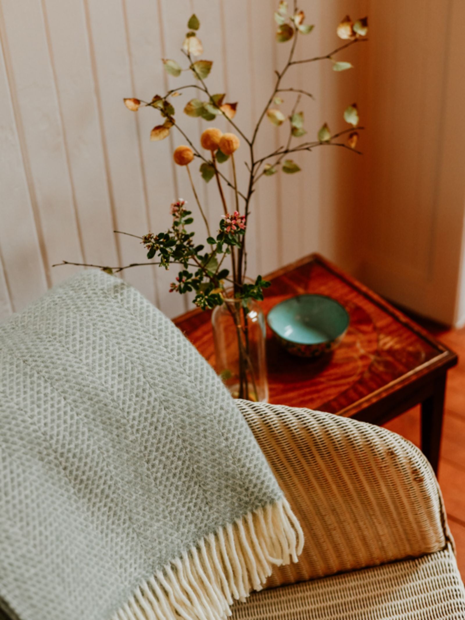 A cozy woven chair draped with a Campbell's of Beauly Stipled Twill Blanket sits beside a small wooden table holding a vase with branches and a ceramic bowl in a warmly lit room.