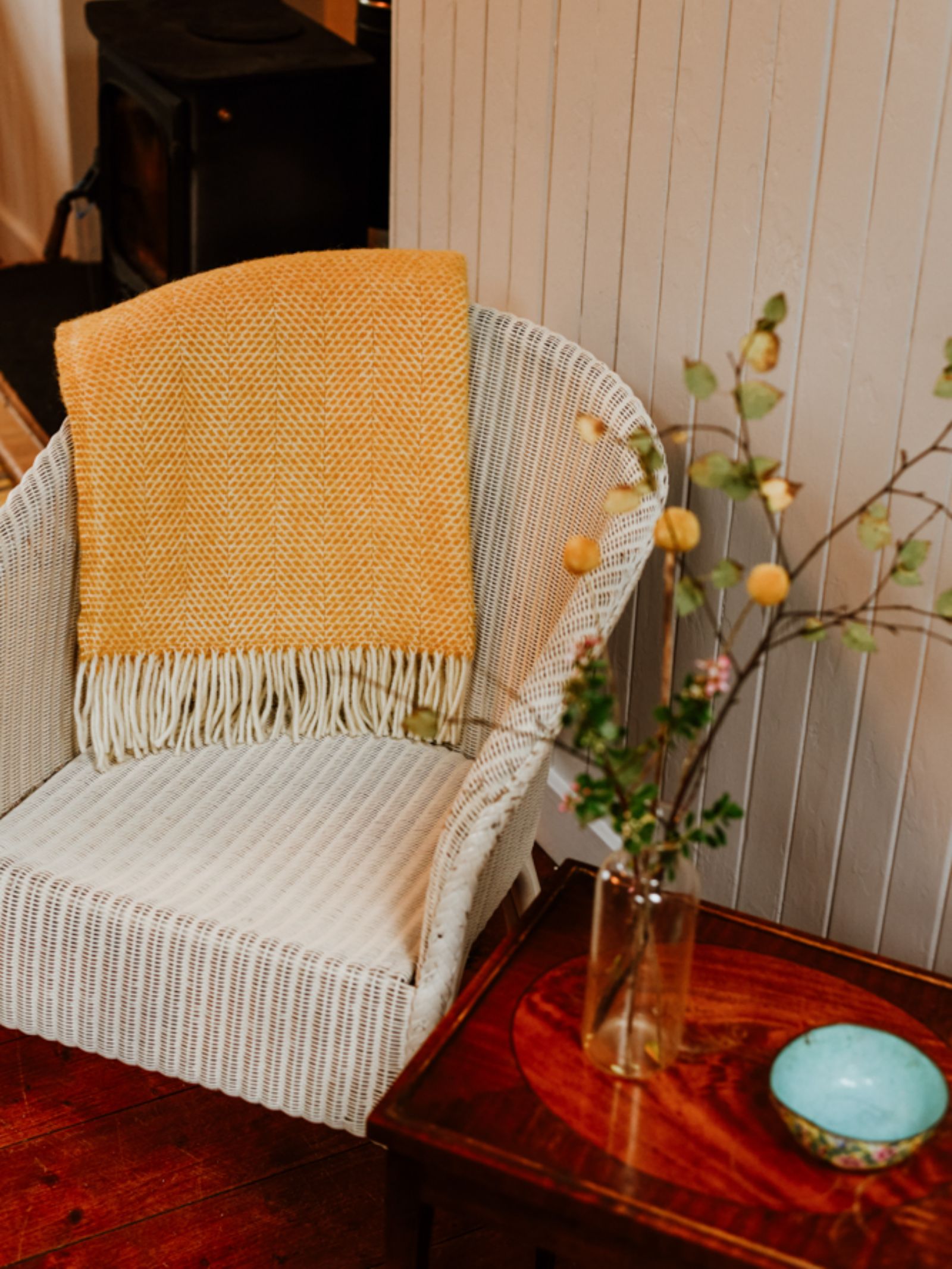 A white wicker chair with a yellow Stipled Twill Blanket from Campbell's of Beauly draped over it sits beside a wooden table holding a vase of branches and a turquoise bowl, with a wood stove in the background.