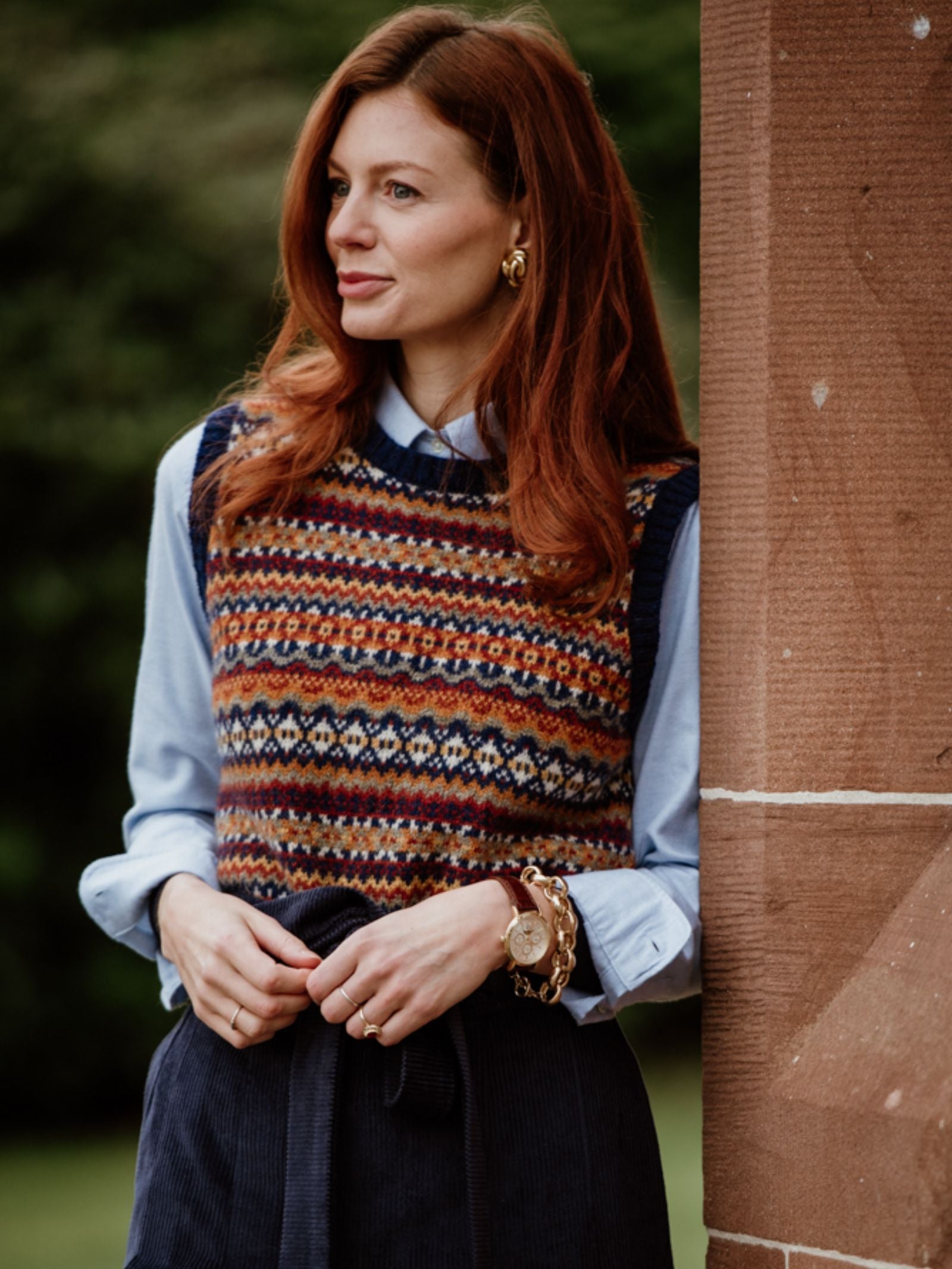 A woman with long red hair stands outdoors by a stone pillar, wearing the Brushed Cotton Shirt from Campbell’s of Beauly under a patterned sweater vest with gold jewelry. She looks to the side, greenery behind—this shirt is a versatile wardrobe staple.