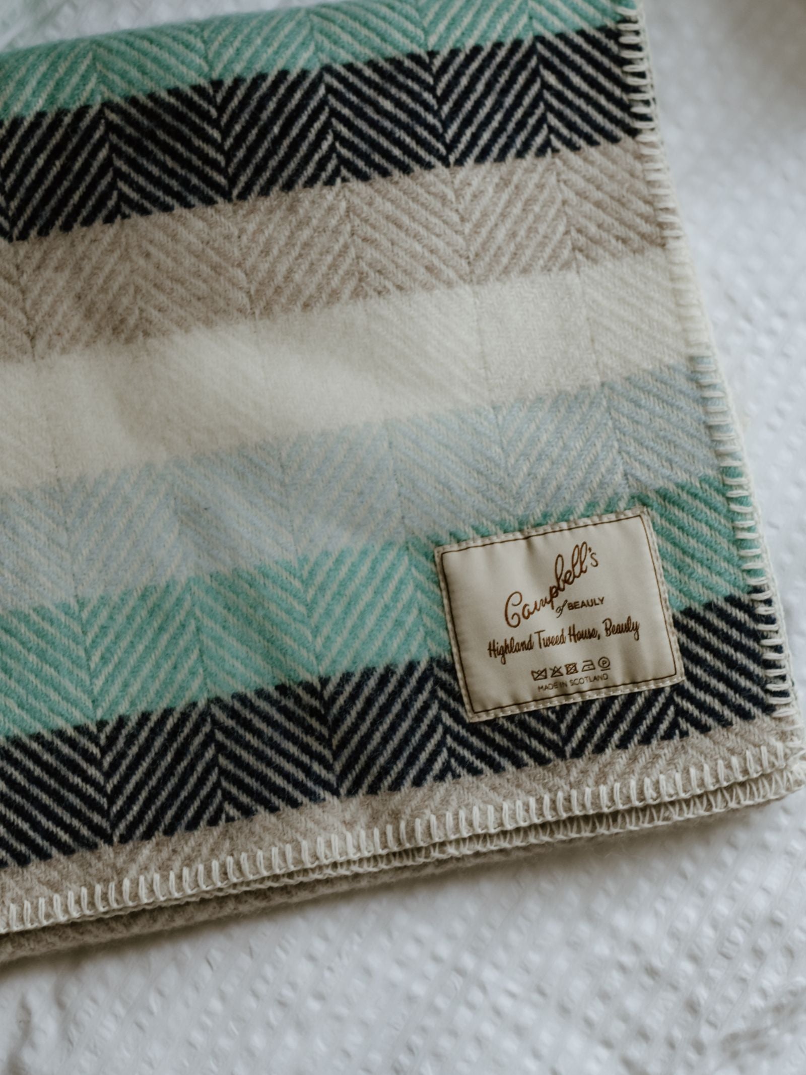 A close-up of Campbell's of Beauly Baby Blanket, showcasing green, black, beige, and cream herringbone wool with a sewn label. The blanket is displayed on a white textured surface.