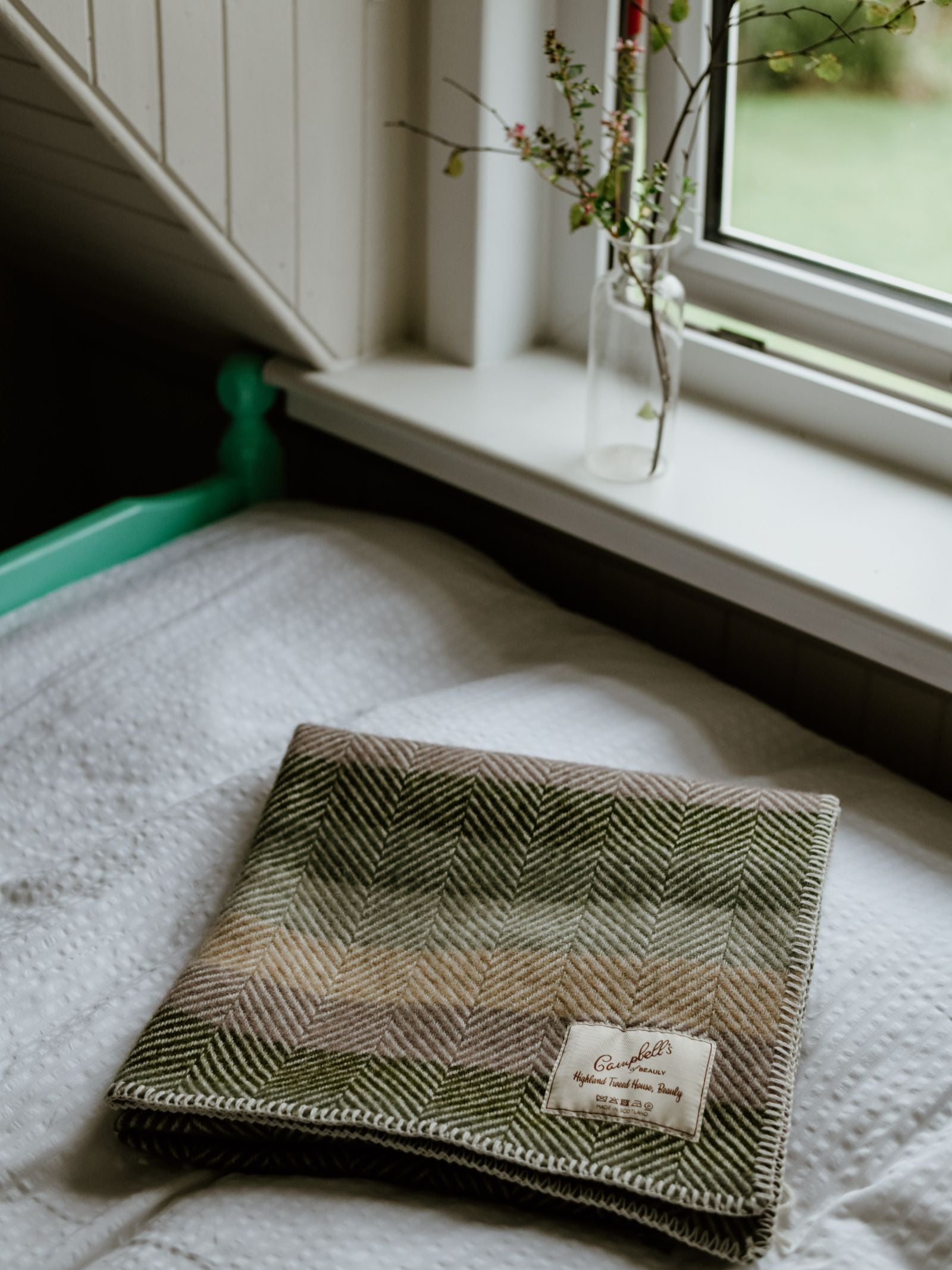 A Campbell's of Beauly Baby Blanket in green and brown herringbone wool is folded on a white bedspread near a window, with a small glass vase holding branches on the windowsill.