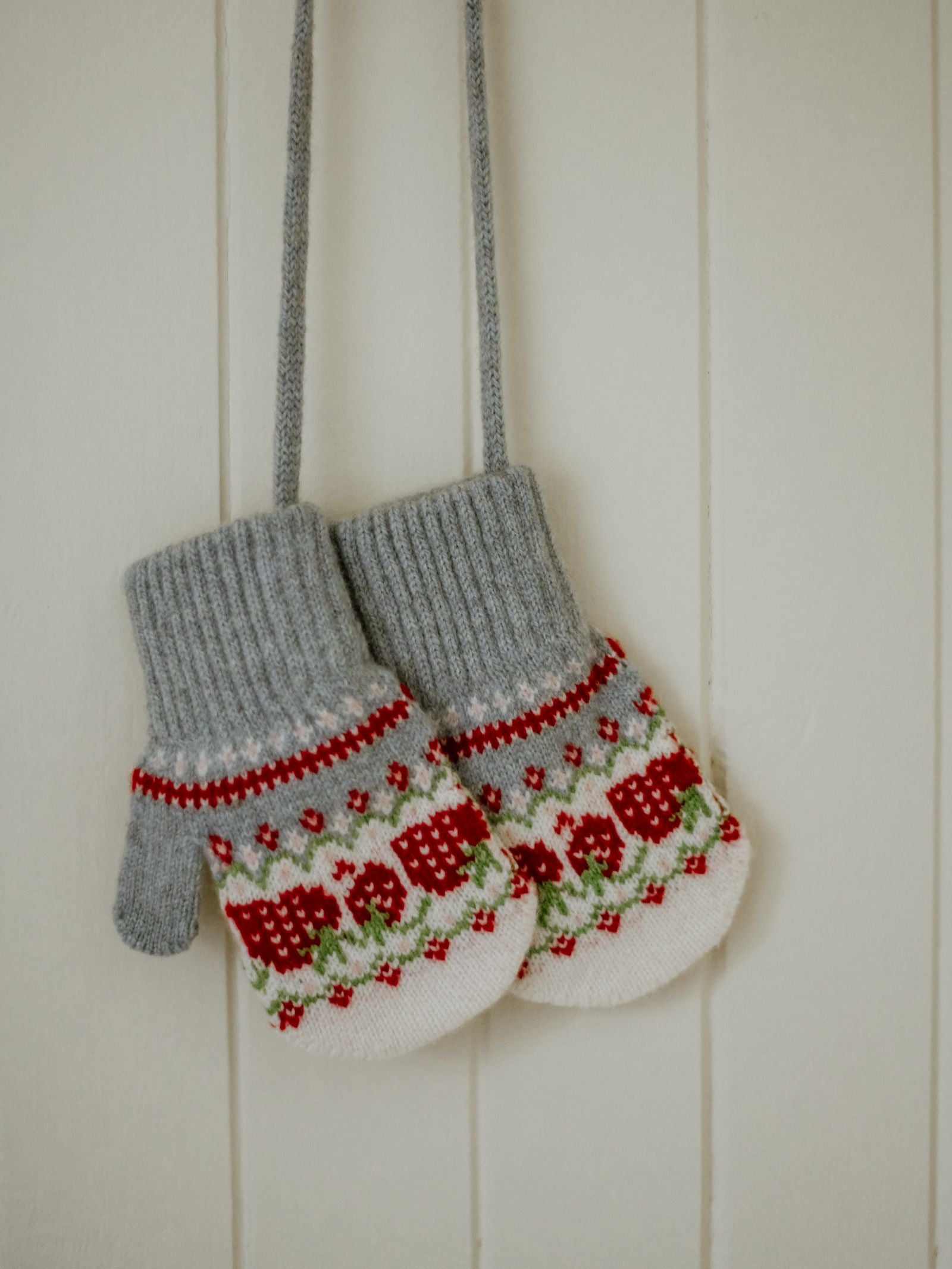 The Mini Lambswool Strawberry Mittens by Campbell's of Beauly, featuring gray cuffs and a red, green, and white pattern, hang by their strings against a white paneled wall.