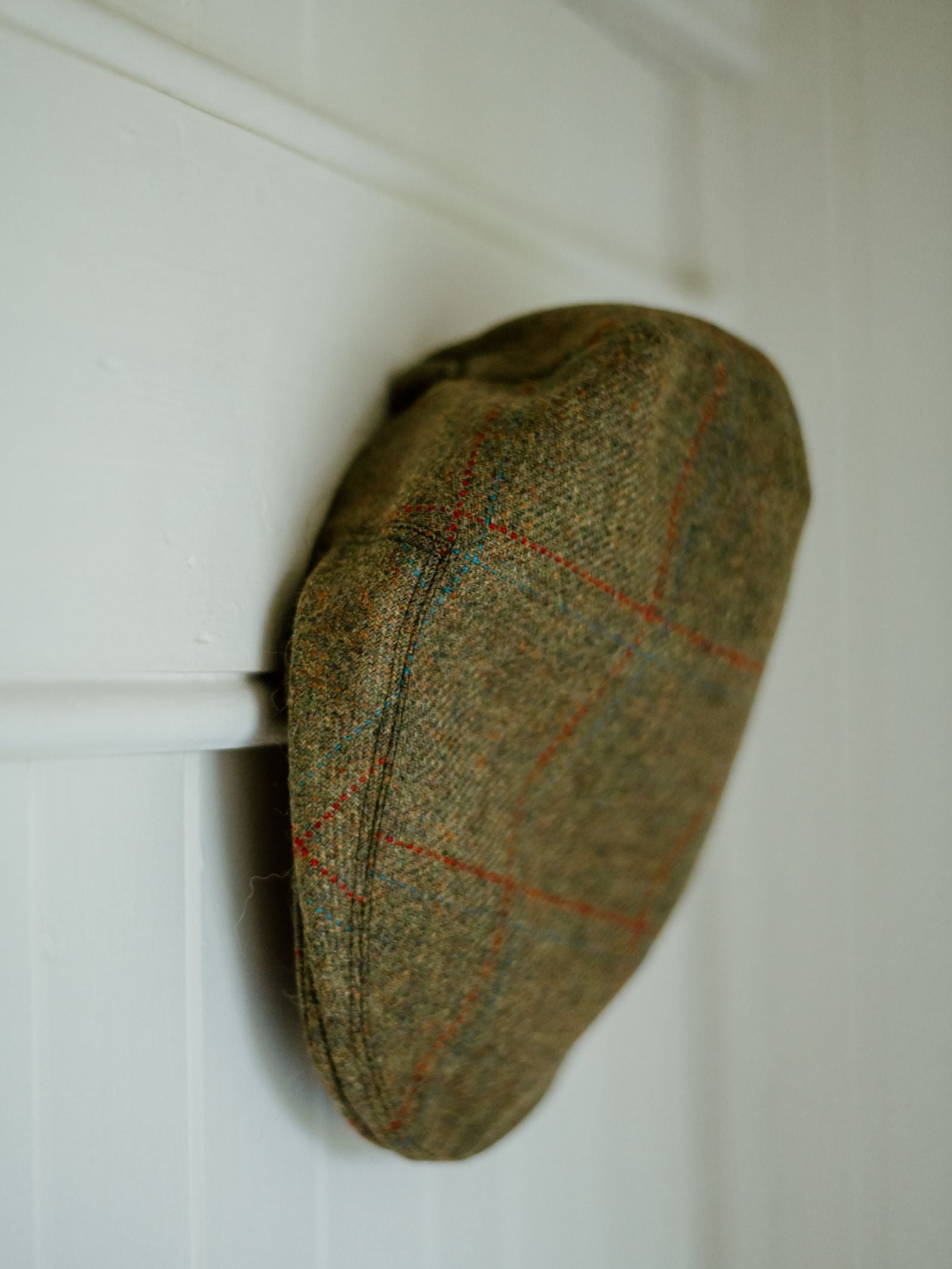 The Mini Tweed Cap by Campbell's of Beauly, featuring a green and brown plaid with red lines, hangs on a white wooden wall, bringing country charm to the space.