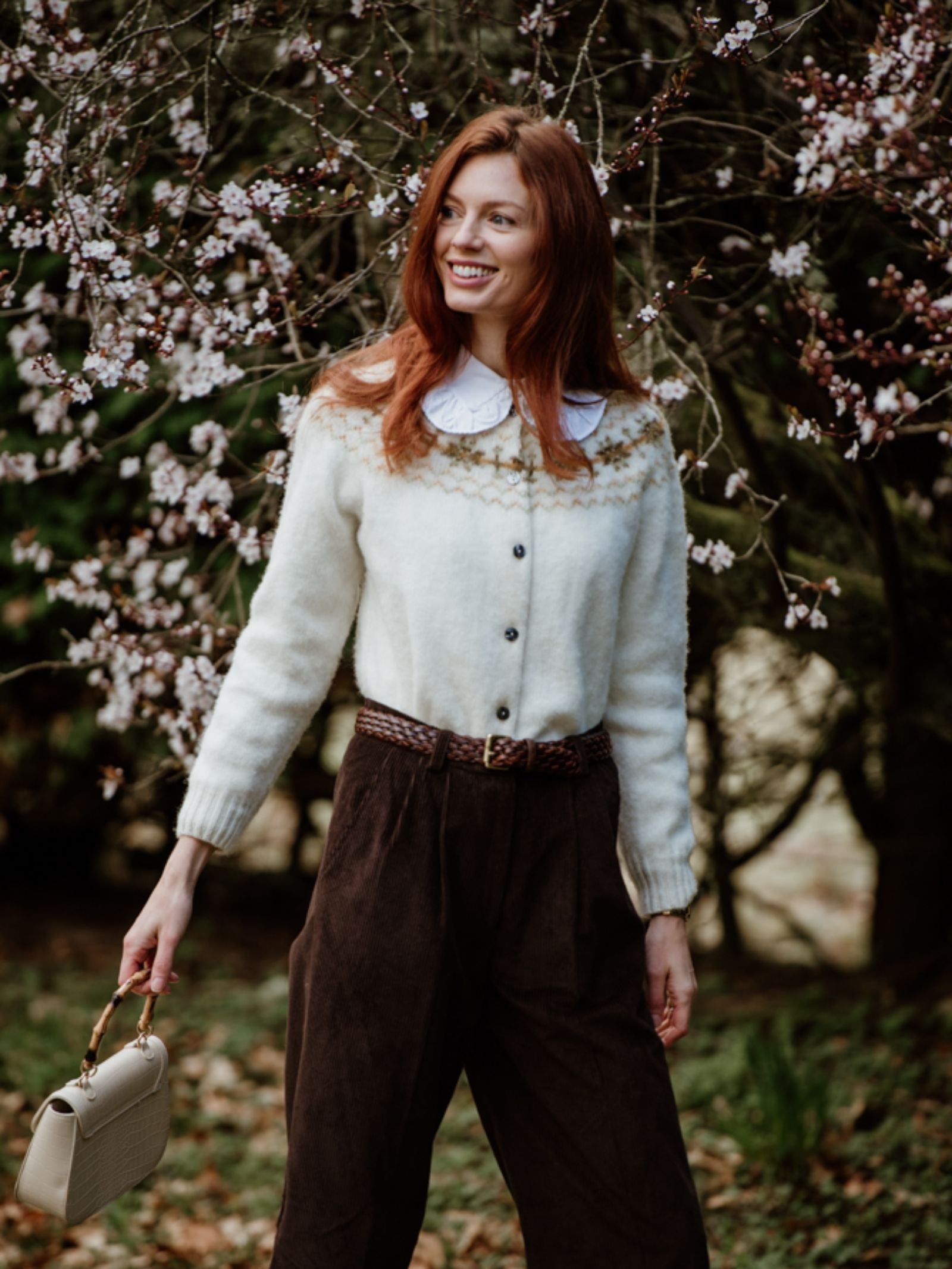 A woman with long red hair smiles outdoors in front of a blossoming tree, wearing the Campbells of Beauly Shetland Fairisle Cardigan, brown pants, and holding a beige handbag by her side.