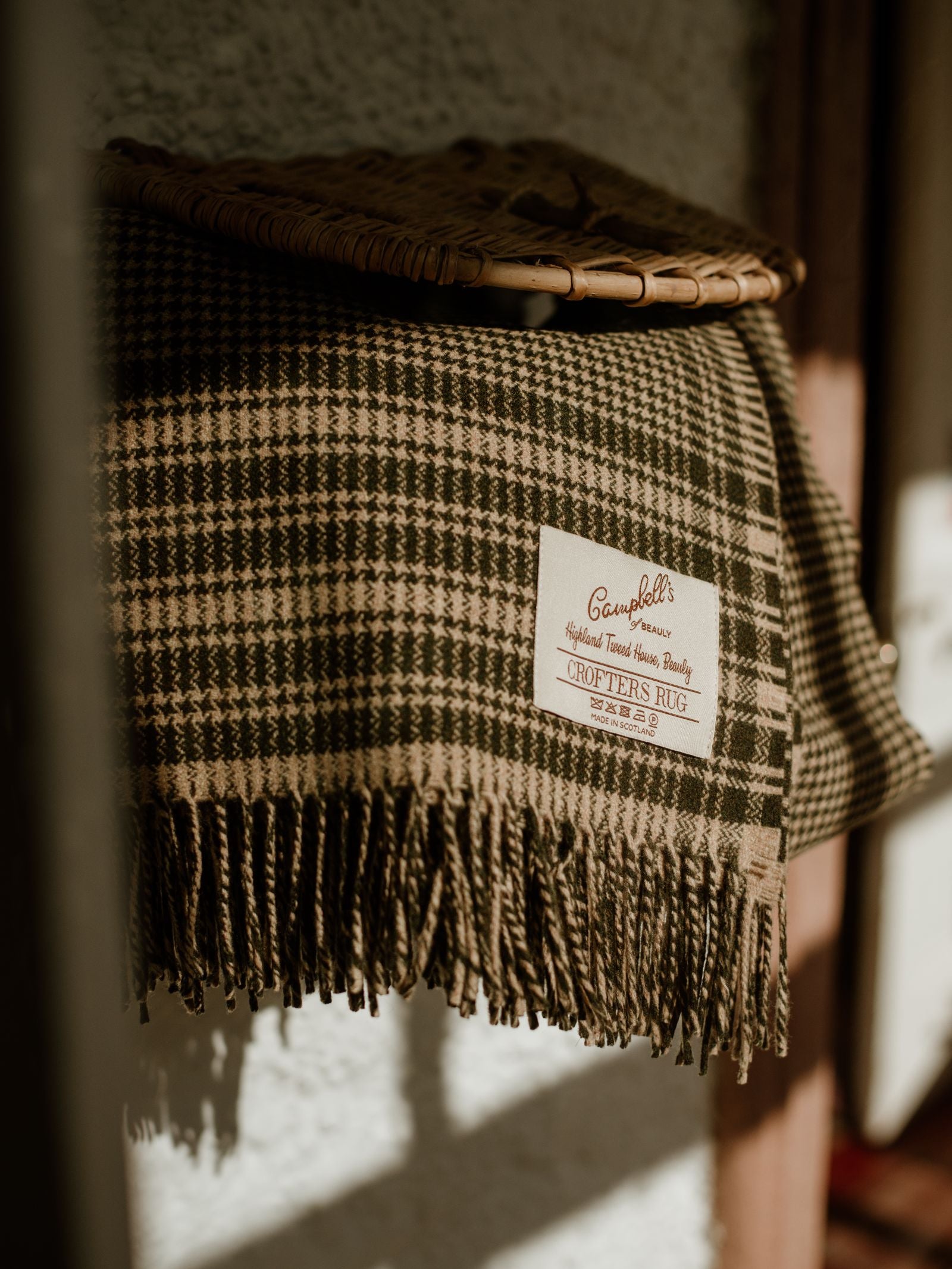 A green and brown Campbell's of Beauly Crofters Blanket with a houndstooth pattern and fringed edges is draped on a surface, evoking a cozy country vibe. A woven basket sits atop it, showing the Campbell's of Beauly label.