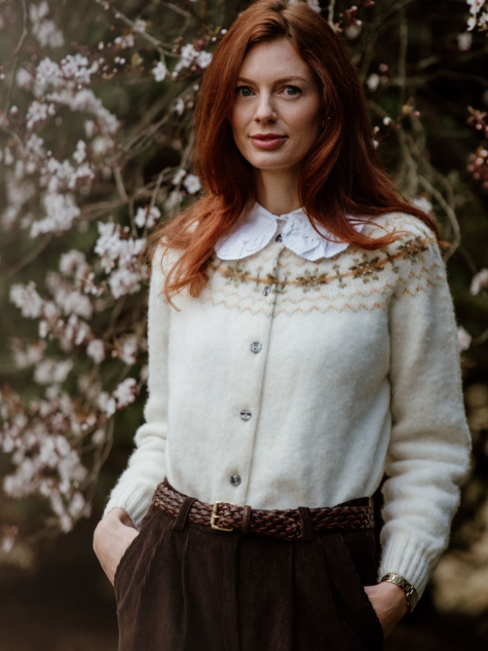 A woman with long red hair stands outdoors before flowering branches, wearing the Campbells of Beauly Shetland Fairisle Cardigan with a decorative pattern and wide collar, paired with dark belted trousers. She looks at the camera, hands in her pockets.