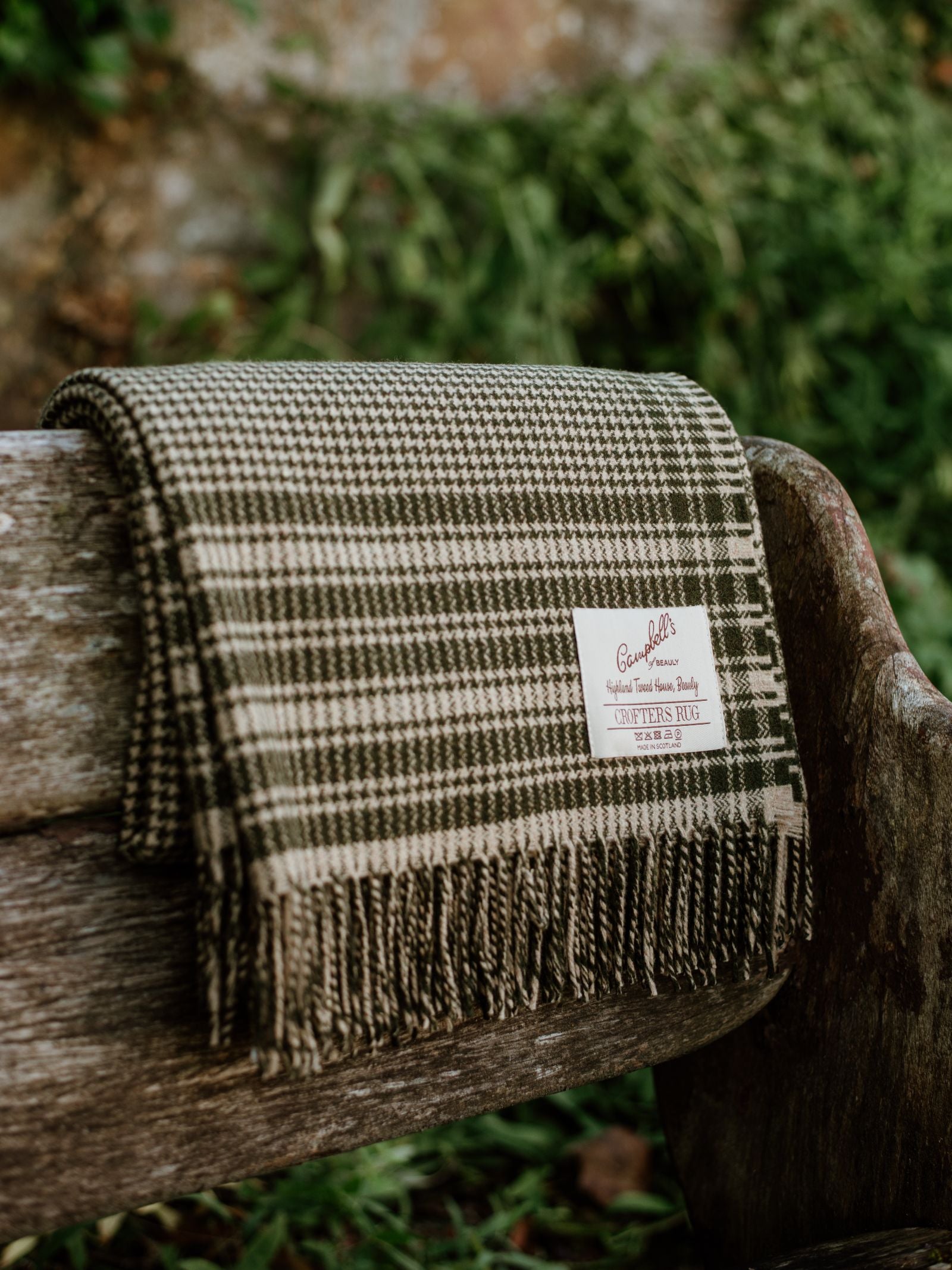 A Crofters Blanket by Campbell's of Beauly, featuring a classic houndstooth pattern and fringed edges, is draped over a rustic bench outdoors, surrounded by lush green grass for a quintessential country look.