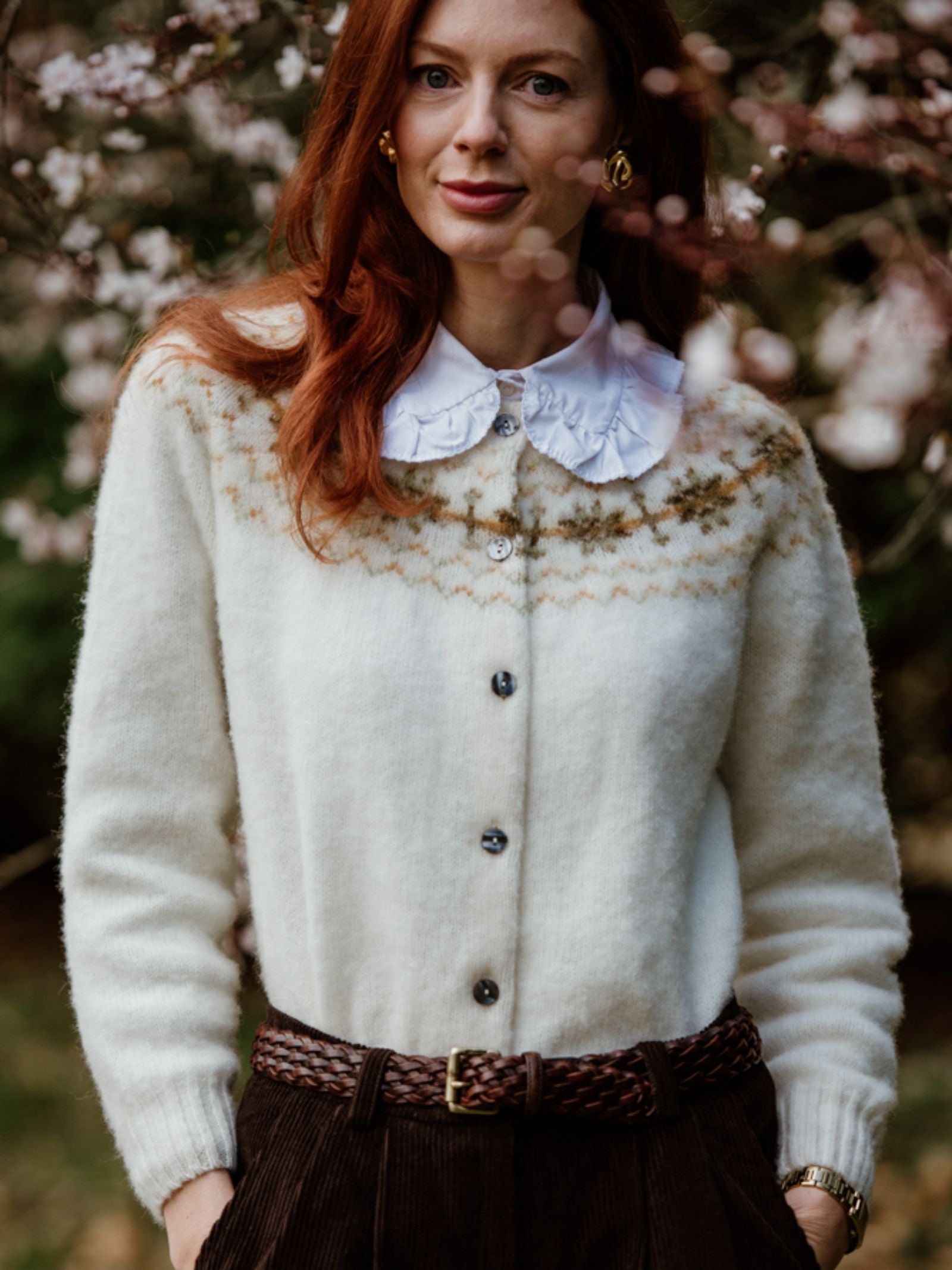 A woman with long red hair stands outdoors before flowering branches, wearing a white Campbell’s of Beauly Shetland Fairisle Cardigan over a collared blouse and high-waisted pants, showcasing classic Scottish knitwear.