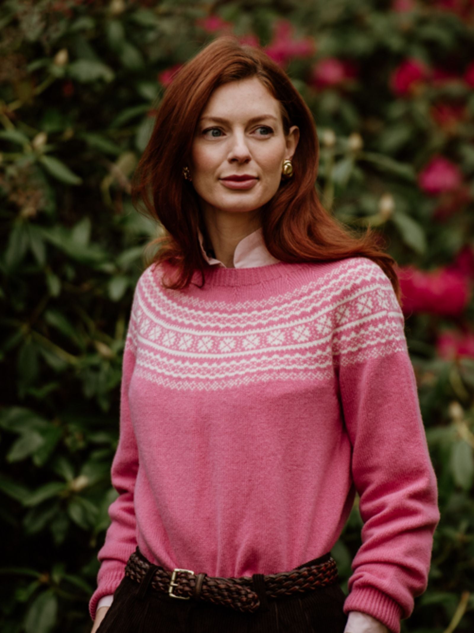 A woman with long red hair stands outdoors amid greenery and pink flowers, wearing a Campbell’s of Beauly Two-Colour Fairisle Yoke Jumper over a collared shirt, paired with dark pants, a braided belt, and gold earrings.