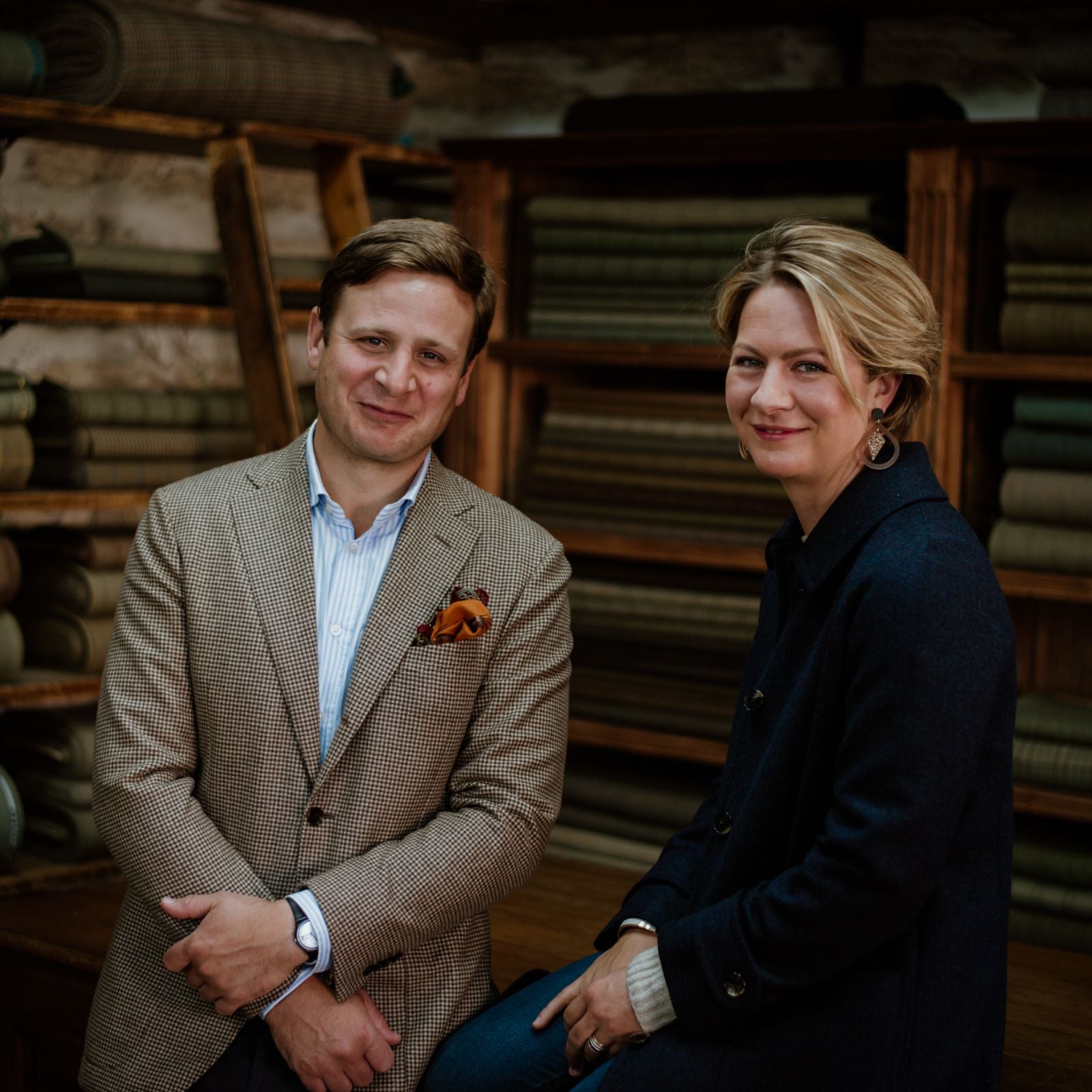 A man in a checked blazer and a woman in a dark coat sit and smile in front of shelves filled with folded fabrics in a warmly lit room.