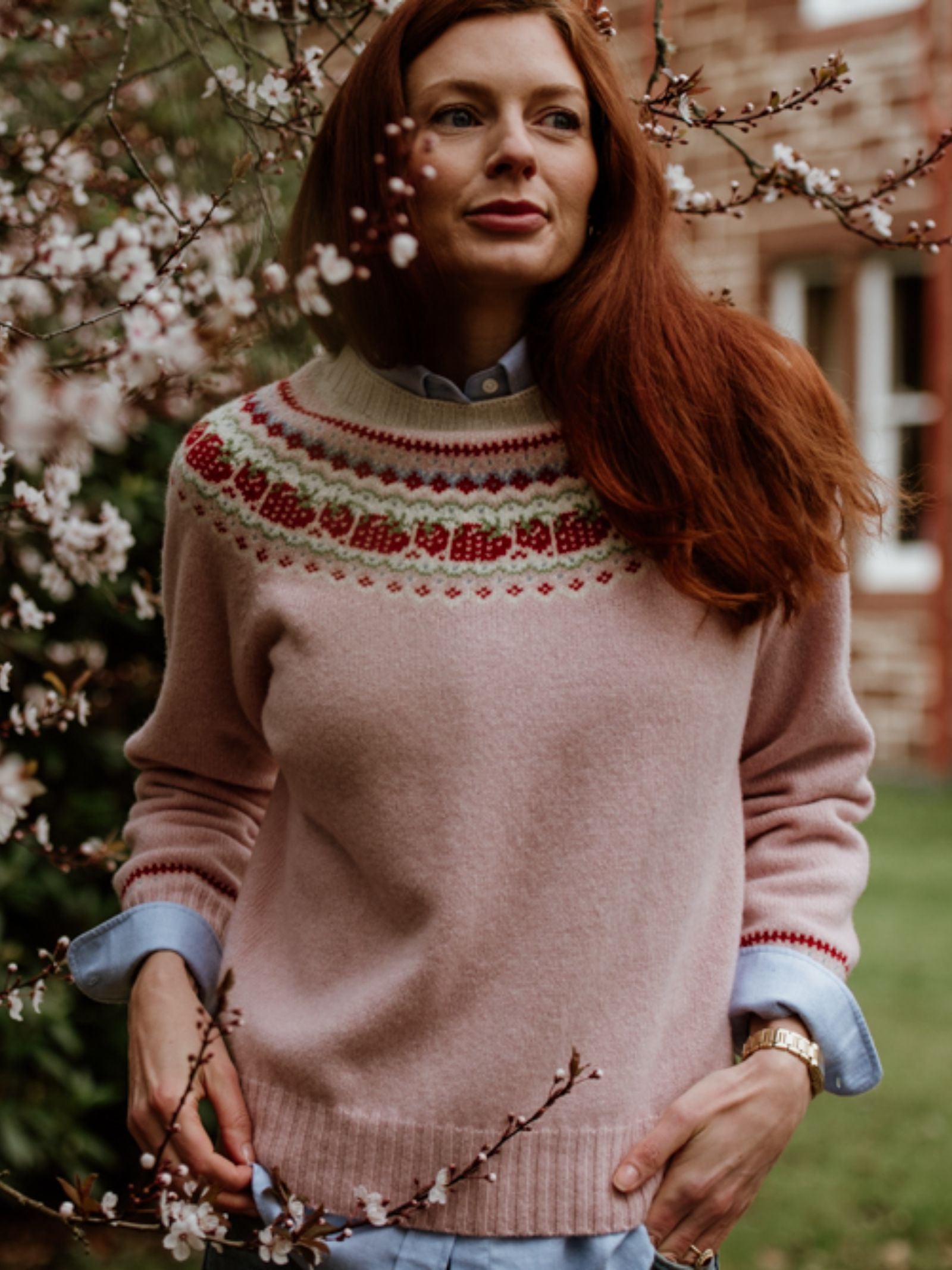A woman with long red hair stands outdoors among blooming branches, wearing the Campbells of Beauly Geelong Lambswool Strawberry Yoke Jumper, with a brick building and Aberdeenshire countryside greenery in the background.