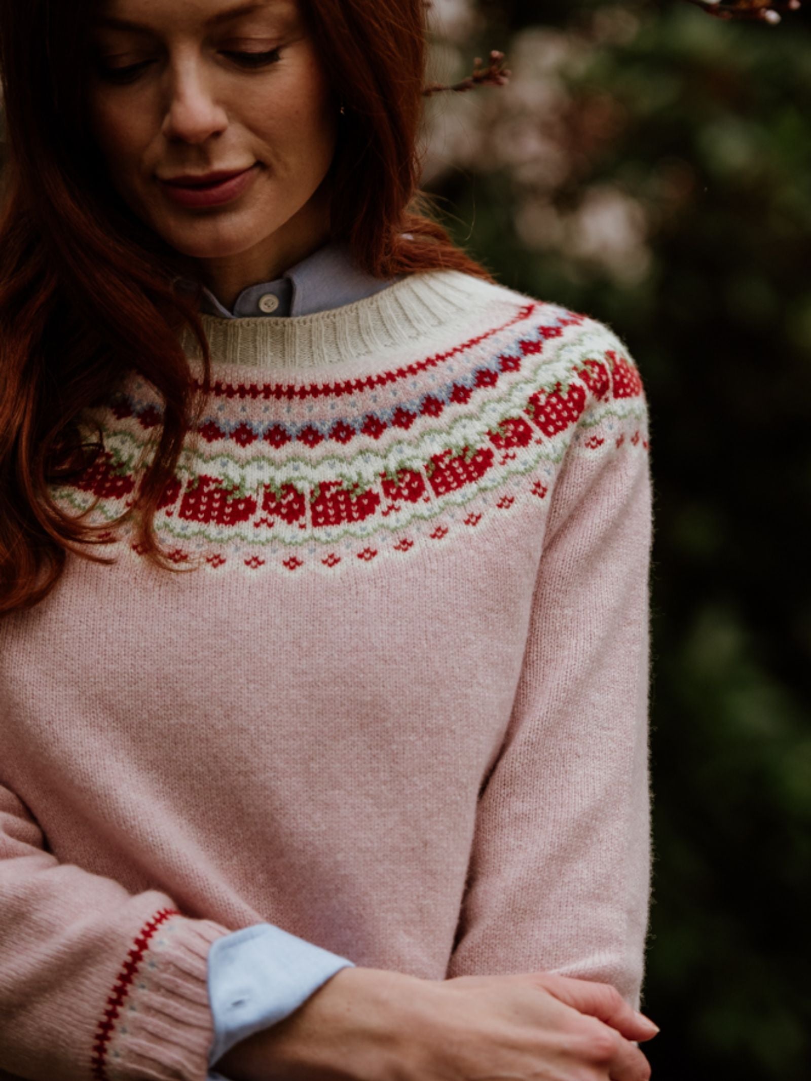 A woman with long red hair wears the Campbells of Beauly Geelong Lambswool Strawberry Yoke Jumper over a pale blue collared shirt, standing outdoors amid blurred Aberdeenshire greenery.