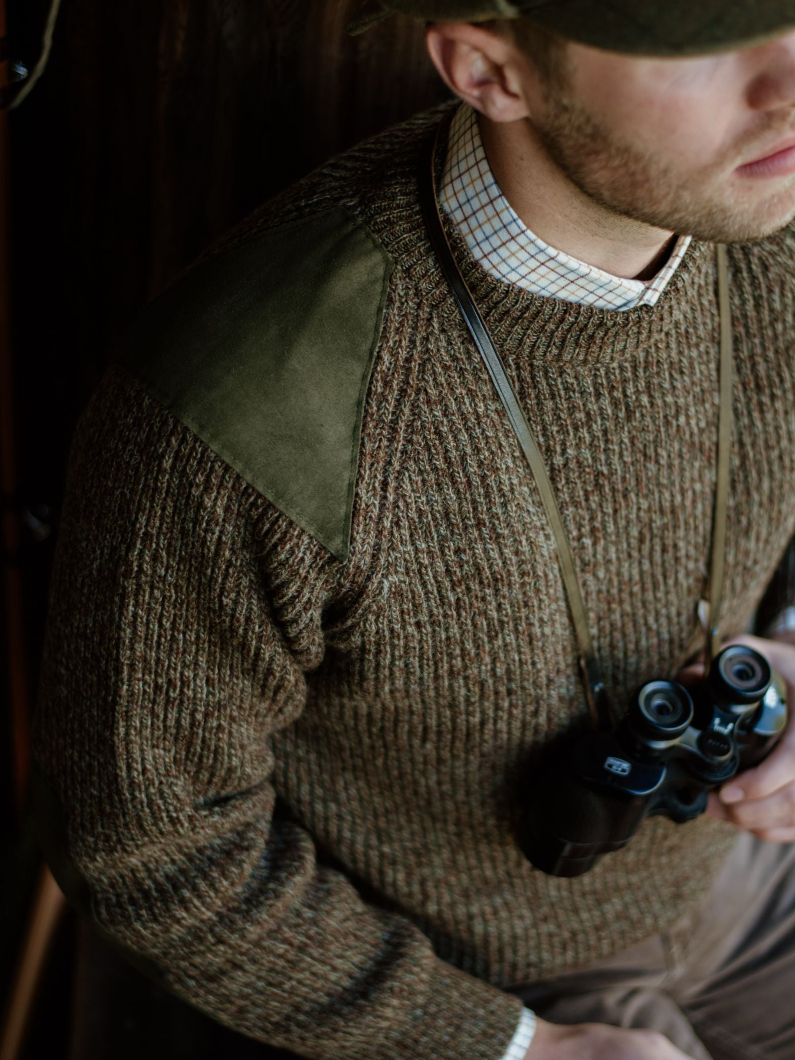 A man wears Campbells of Beauly’s Scotia Suede Jumper in olive with green suede shoulder patches, a plaid collared shirt underneath, and a green cap. Binoculars hang from his neck; his face is partly out of frame.