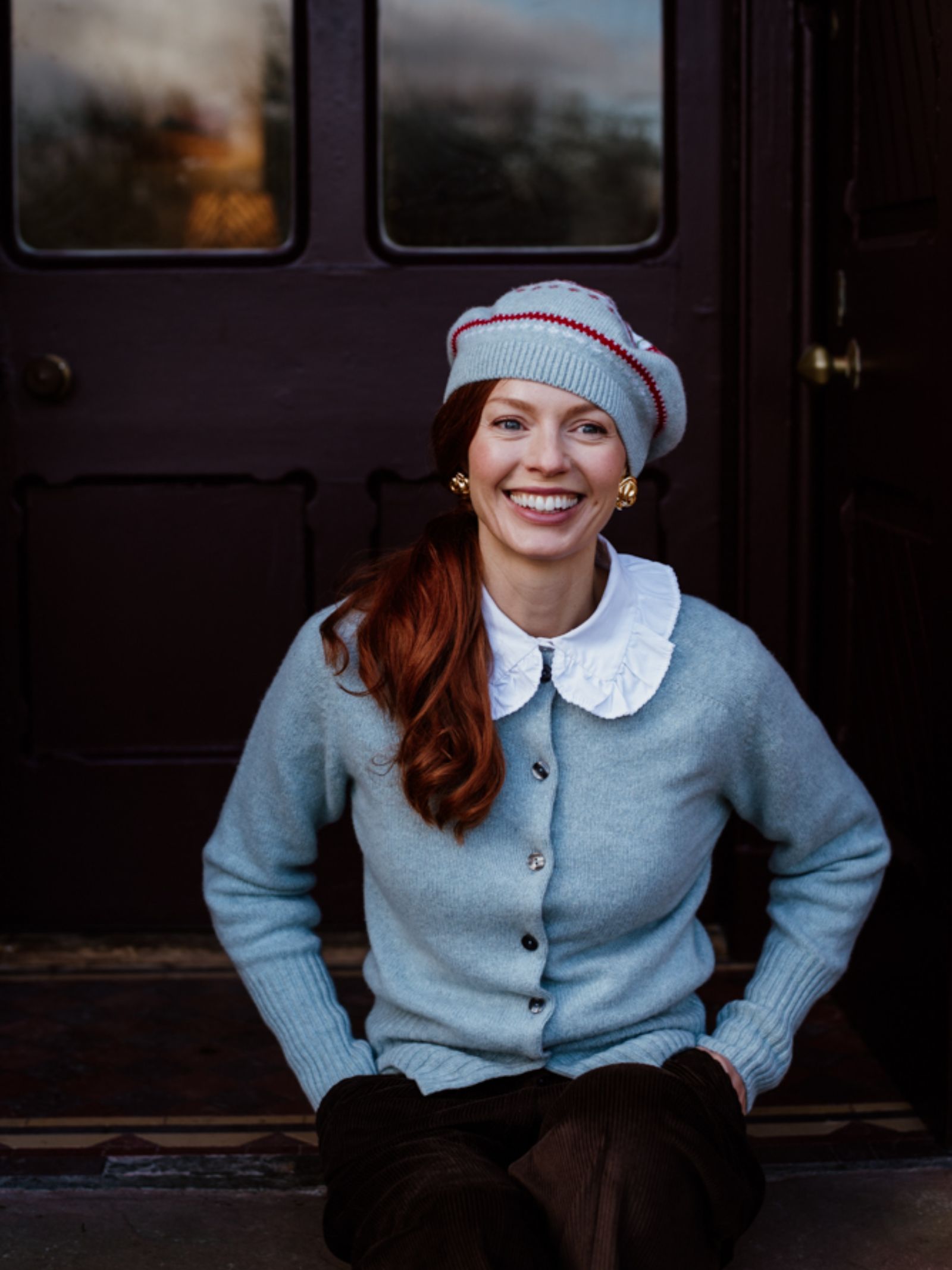 A smiling woman sits in front of a dark doorway, wearing a Campbells of Beauly Strawberry Beret, a light blue cardigan over a white ruffled blouse, brown pants, and gold earrings. Her hands rest casually on her knees.