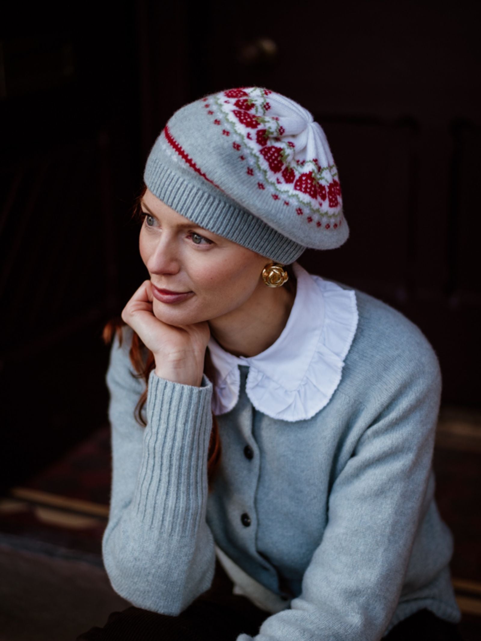 A woman with red hair wears a light gray sweater over a white collared shirt and the Strawberry Beret by Campbells of Beauly, which features red and white patterns. Seated, she rests her chin on one hand and smiles softly while gazing to the side.