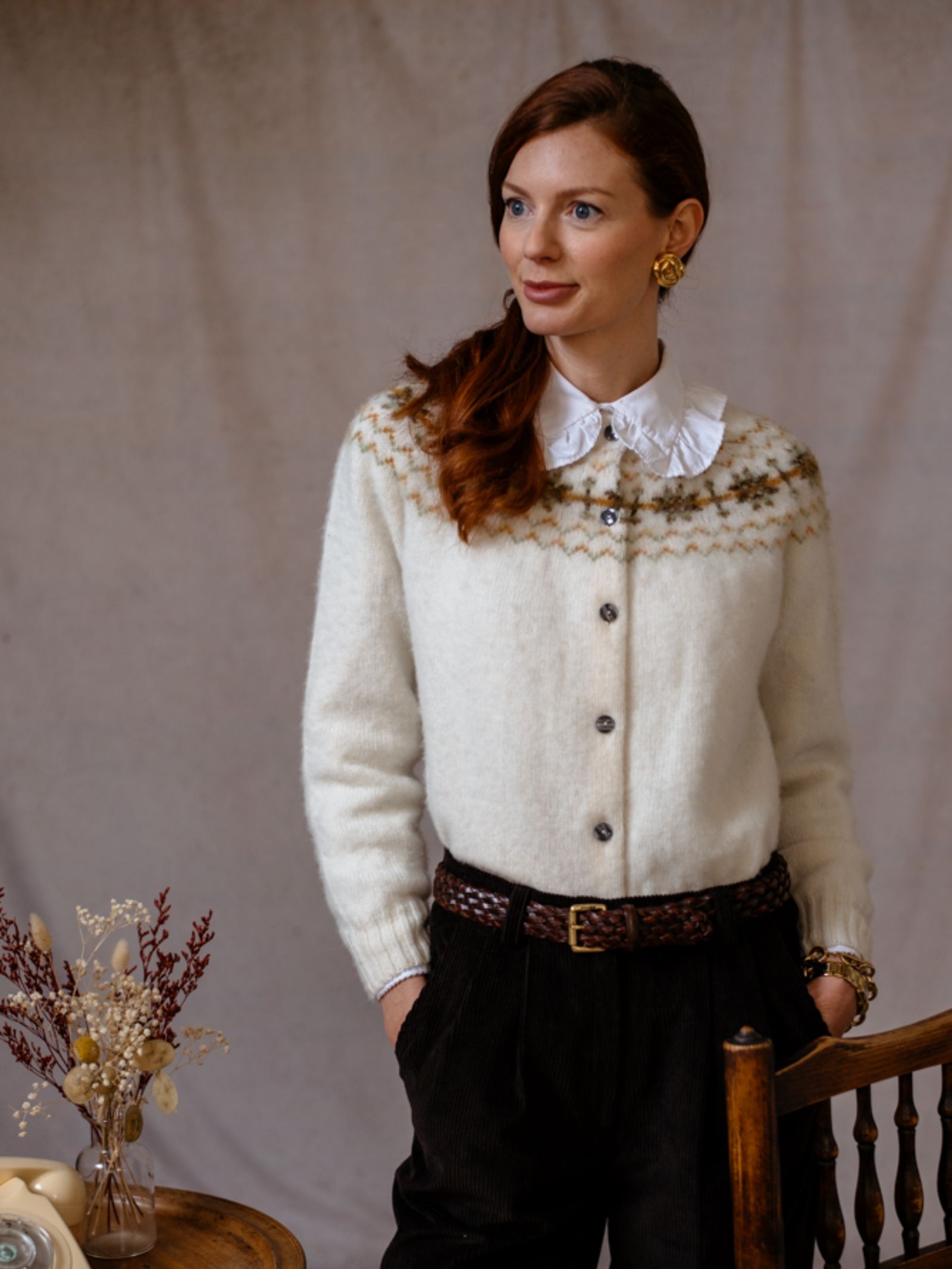 A woman with long brown hair, wearing the Campbells of Beauly Shetland Fairisle Cardigan over a collared shirt, dark pants, and a brown belt, stands indoors beside a wooden chair and a table with dried flowers.