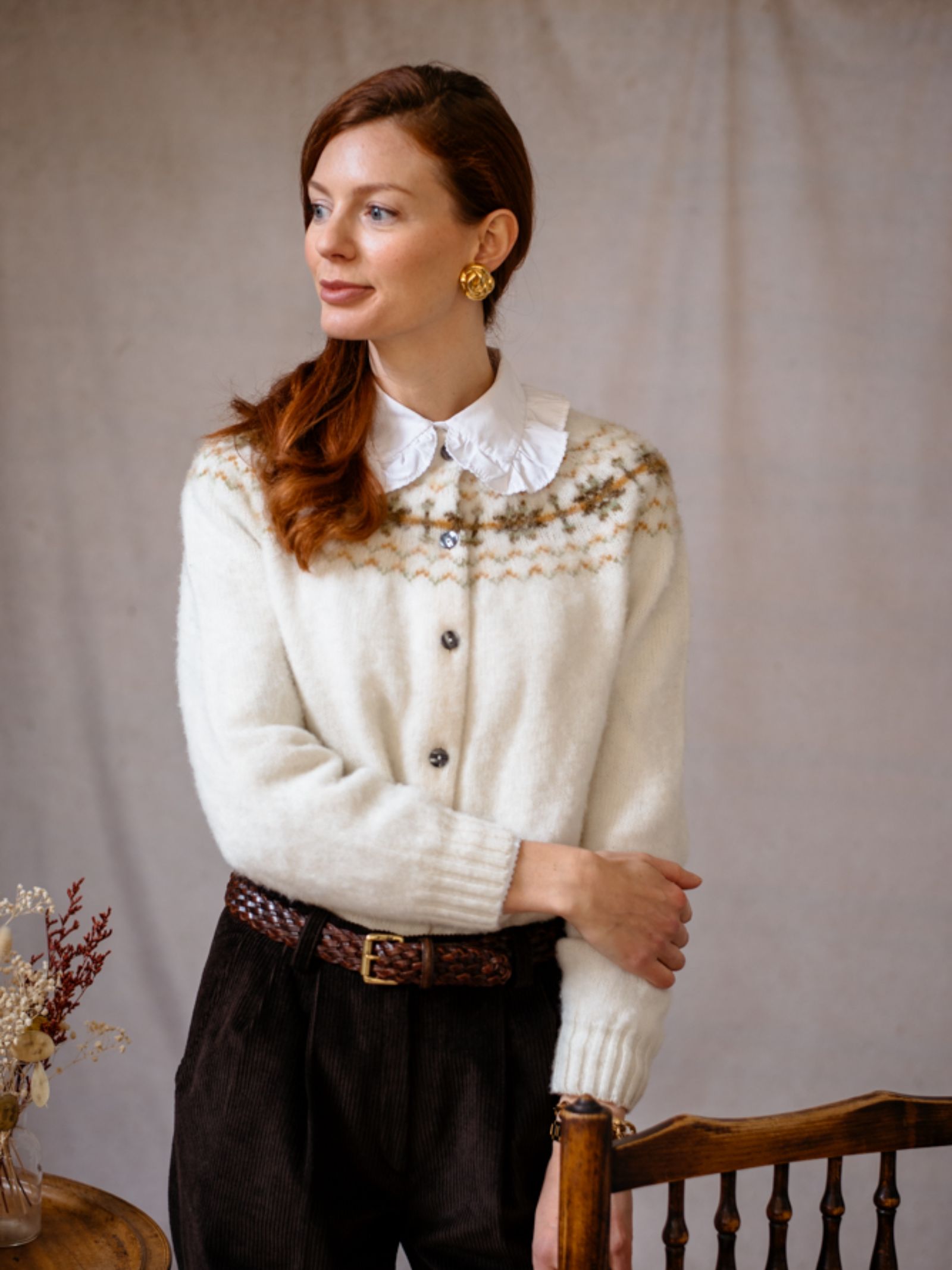 A woman with long red hair, wearing a Campbells of Beauly Shetland Fairisle Cardigan over a collared shirt, brown trousers, and gold earrings, stands indoors by a wooden chair, looking to the side.