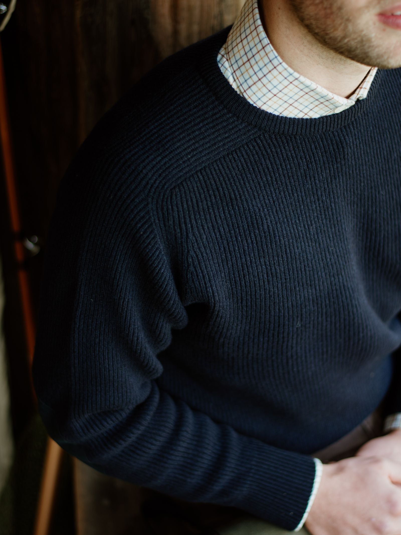A person wearing the Campbells of Beauly Fishermans Rib Crew Jumper in navy blue over a white shirt with a blue and red check pattern, seated indoors with their face partially out of frame.