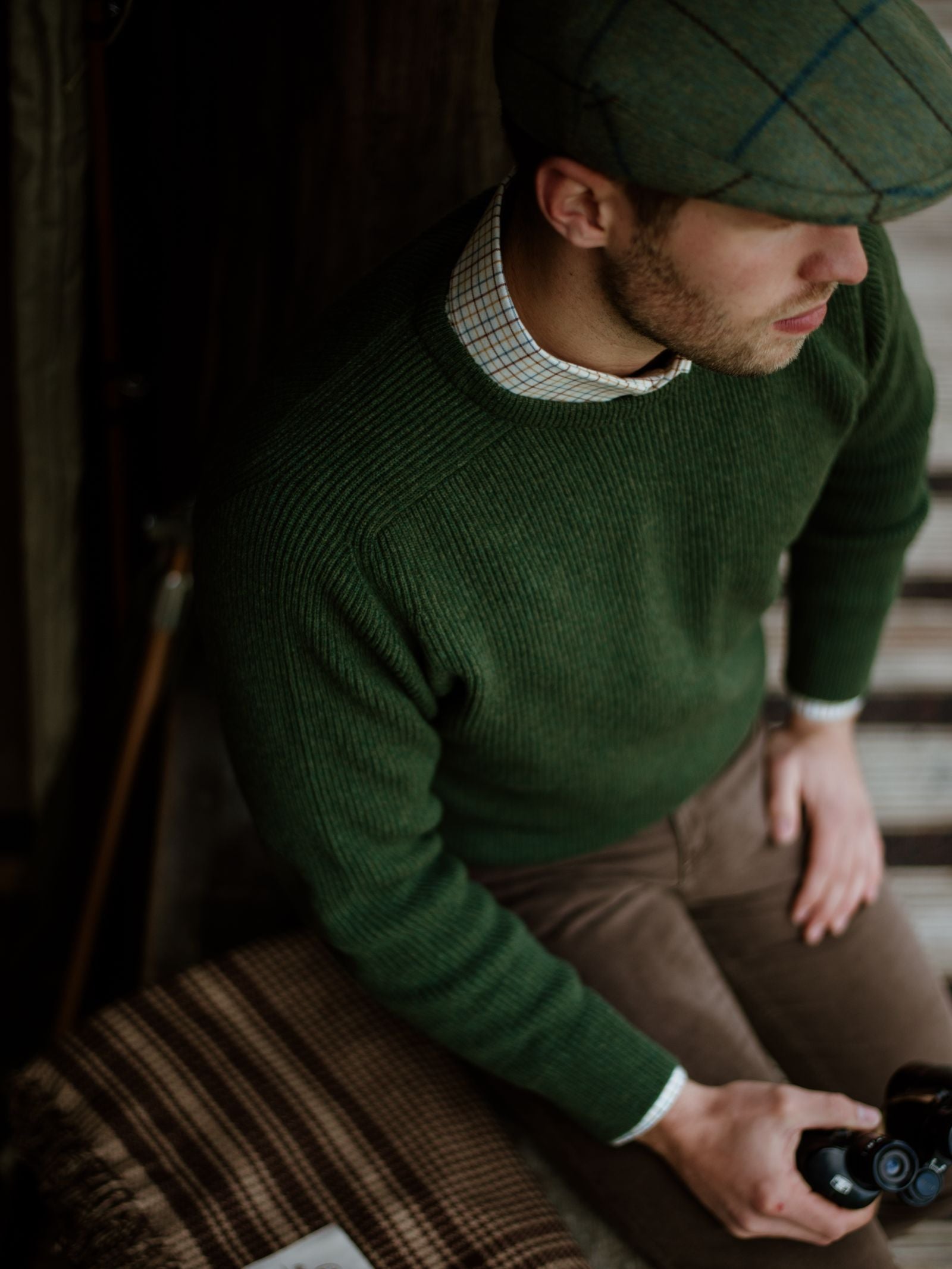 A man in a Campbells of Beauly Fishermans Rib Crew Jumper, brown pants, and a green plaid cap sits indoors holding an object beside a plaid wool blanket, gazing to the side.