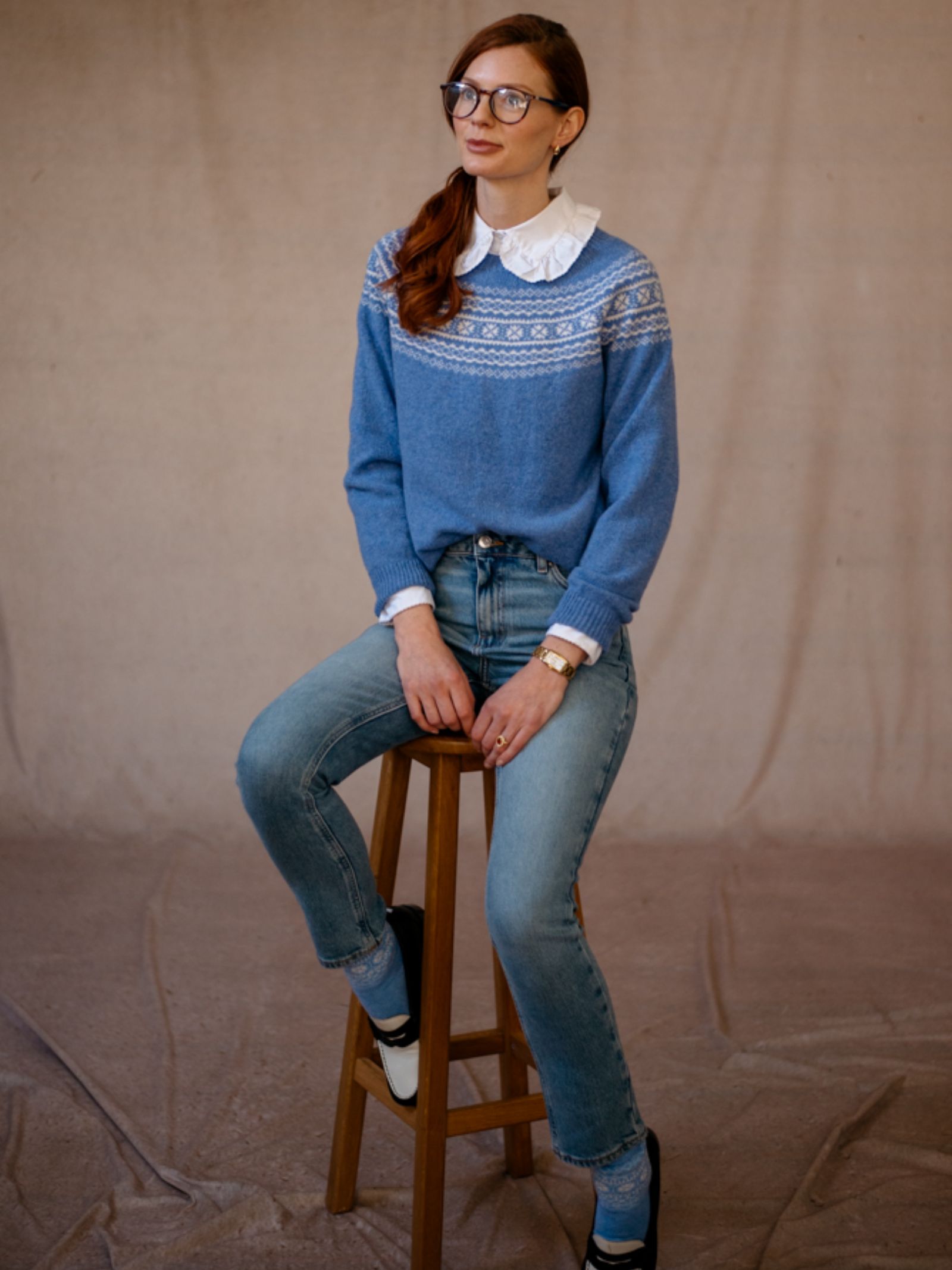 A woman with long brown hair and glasses sits on a wooden stool, wearing Campbells of Beauly Two-Colour Fairisle Yoke Jumper over a white blouse, blue jeans, and black-and-white shoes, against a simple beige backdrop.