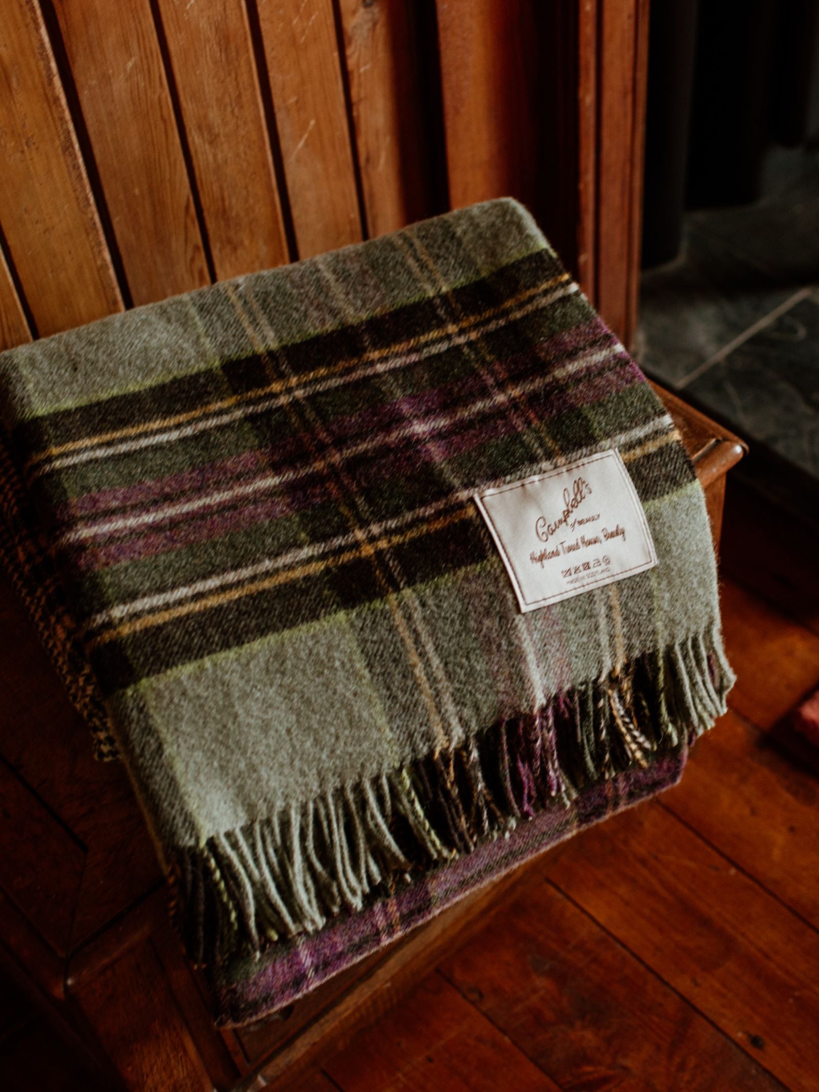 The Strathglass Throw by Campbell's of Beauly, a folded plaid lambswool blanket in green, purple, and brown stripes, rests on a wooden bench. A white label accents the corner, completing this cosy Yorkshire setting.