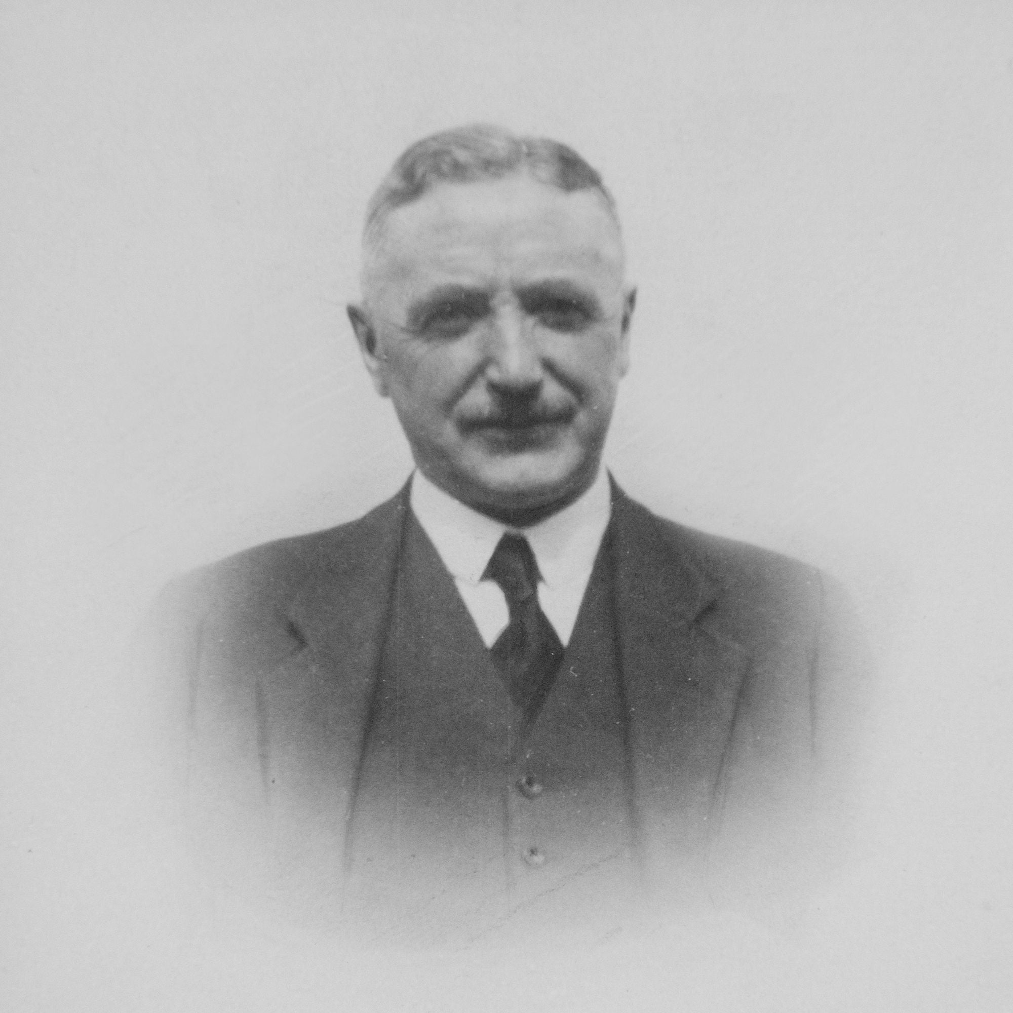 Black and white portrait of an older man with short hair and a mustache, wearing a suit, tie, and vest, looking directly at the camera with a neutral expression.