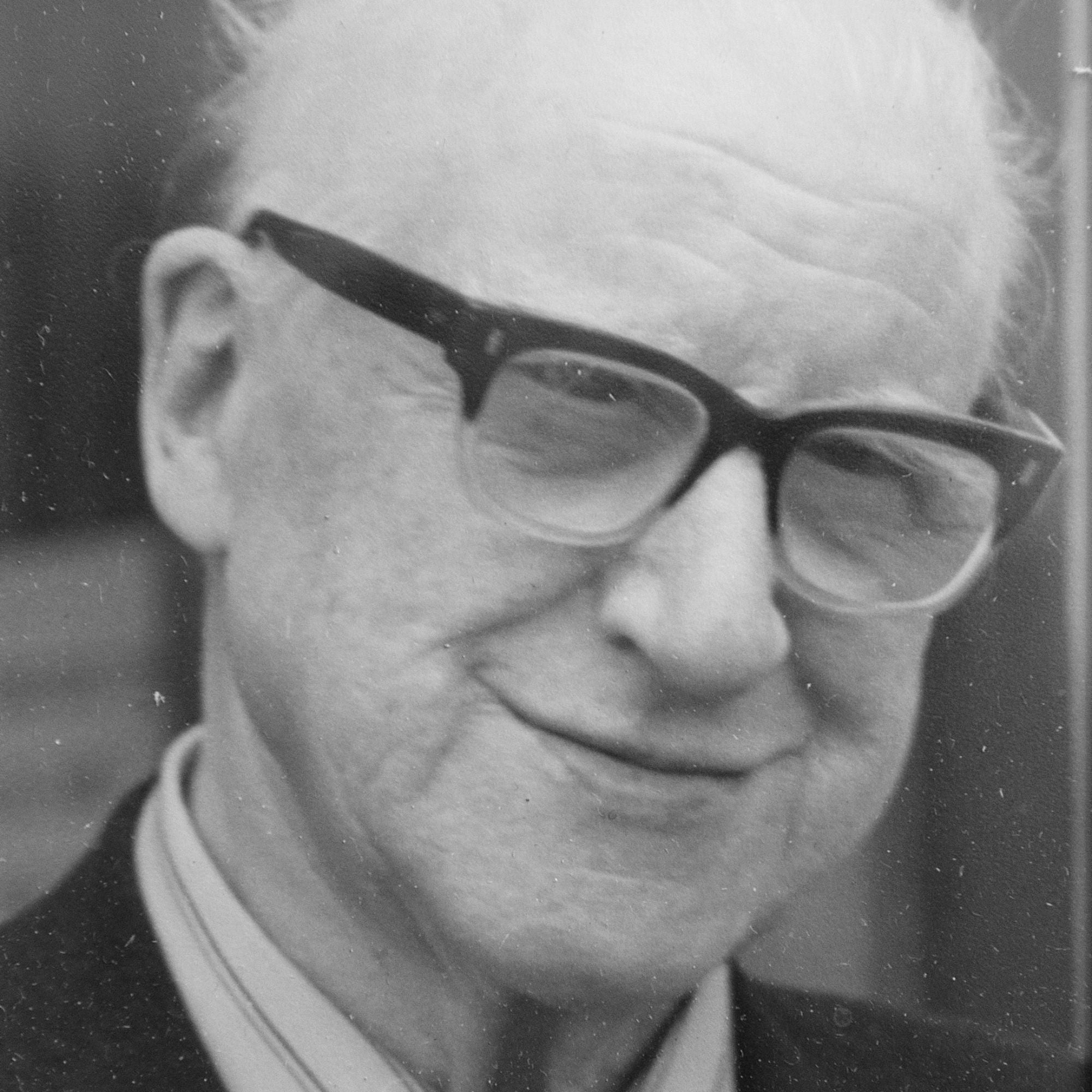 An elderly man with light skin, white hair, and glasses smiles gently. He is wearing a collared shirt and a dark jacket. The photo is black and white and has a close-up focus on his face.