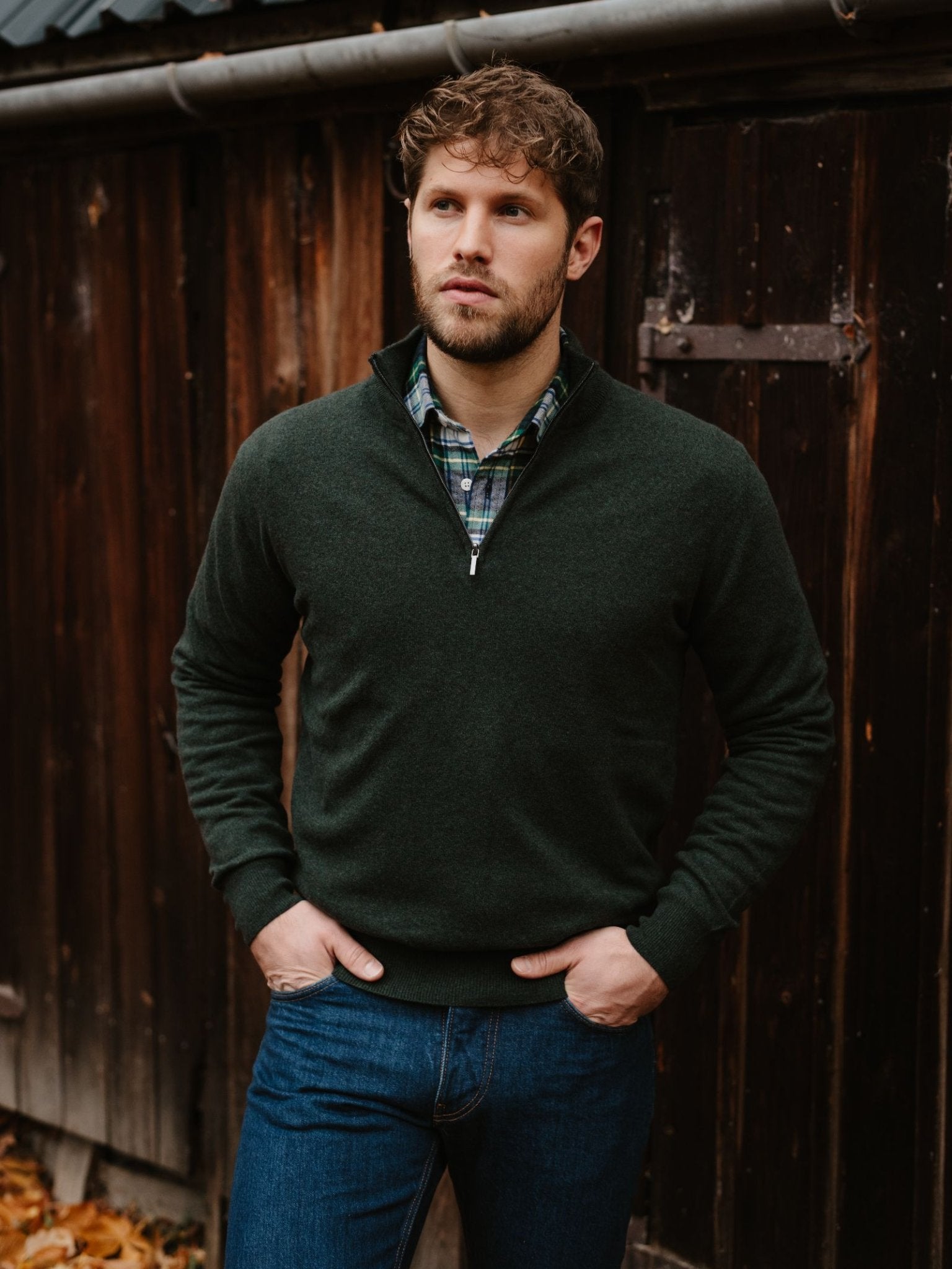 A man with short brown hair and a beard stands before a dark wooden wall, wearing a green Merino & Cashmere Quarter Zip Jumper from Campbell's of Beauly over a plaid shirt and blue jeans, his hands in his pockets.