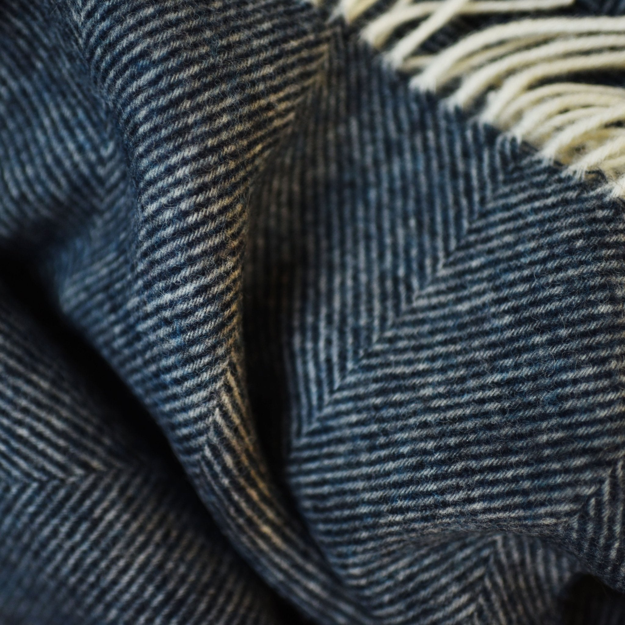 Close-up of a textured, dark blue and white herringbone pattern with soft folds and fringed edges on the Campbells of Beauly Herringbone Lambswool Blanket.