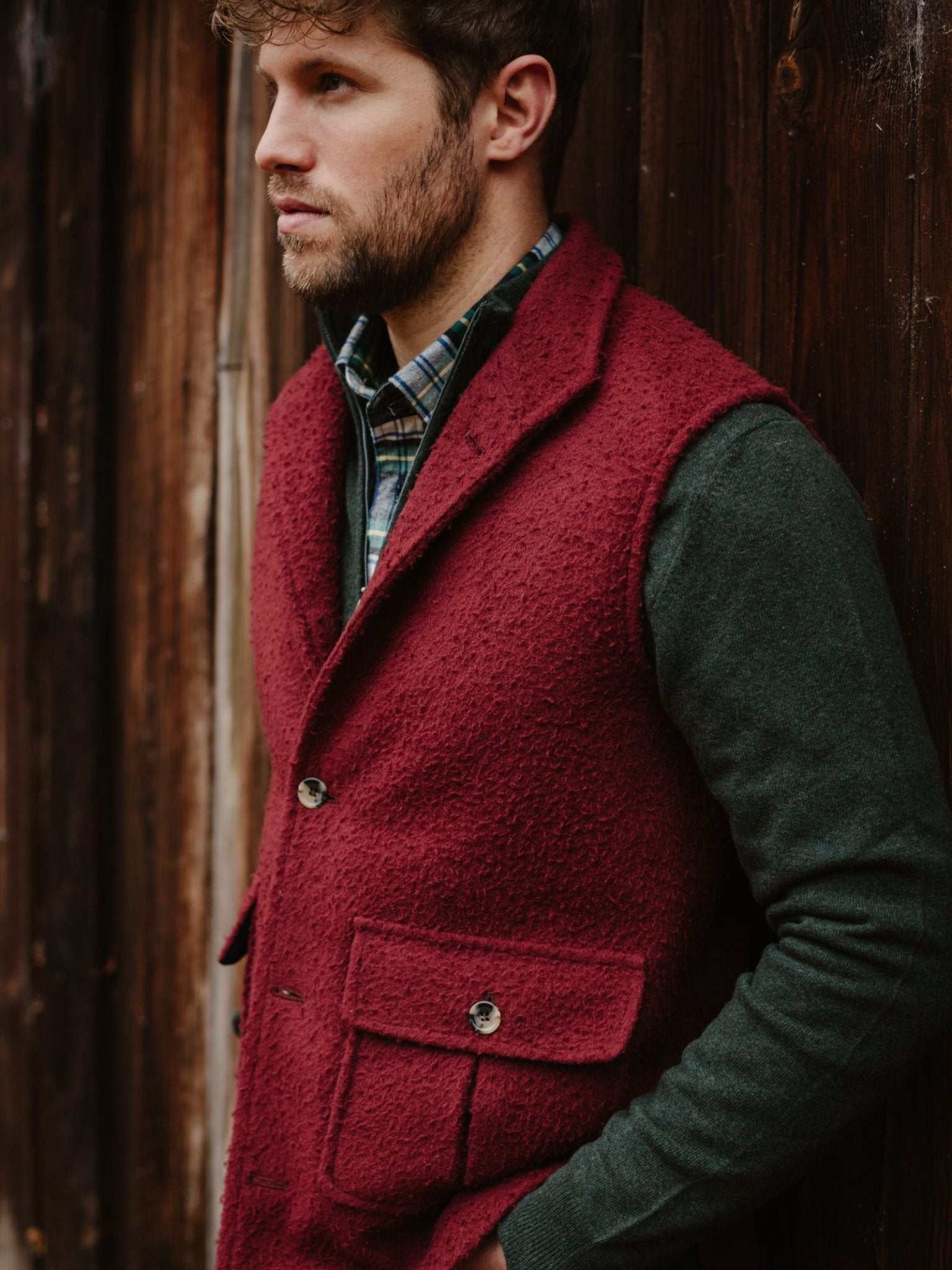 A bearded man, wearing a green shirt and the Casentino Fleece Gilet by Campbell's of Beauly, stands sideways against a wooden wall, gazing into the distance.