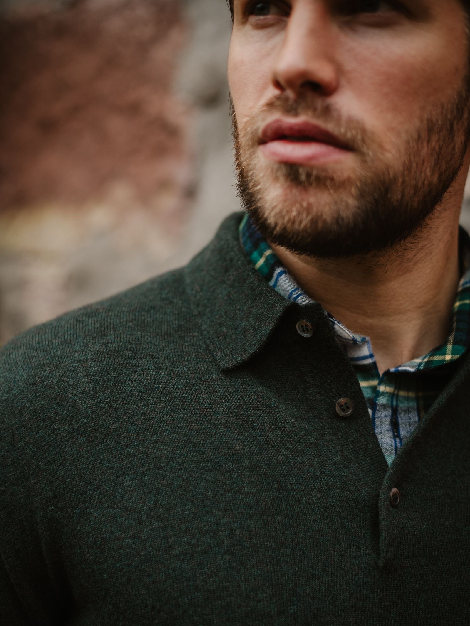 A close-up of a bearded man in refined casual knitwear—a green sweater layered over a Campbell's of Beauly Merino & Cashmere Shirt Jumper with green and blue plaid—looking away from the camera against a blurred background.