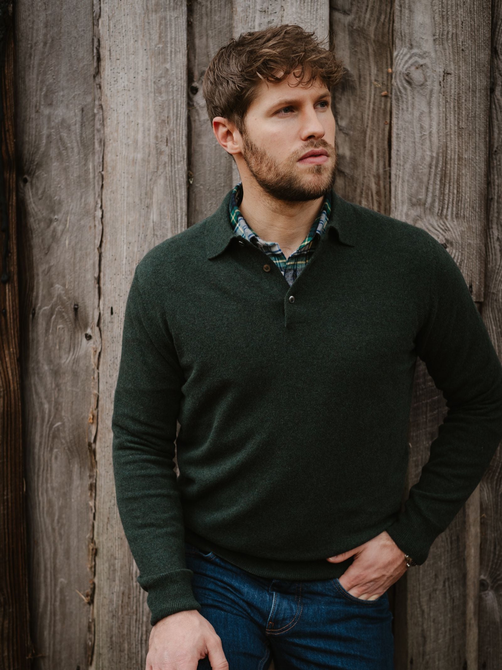 A man with short, wavy brown hair and a beard stands against a rustic wooden wall, wearing Campbell's of Beauly Merino & Cashmere Shirt Jumper layered over a green plaid shirt and blue jeans, gazing thoughtfully into the distance.