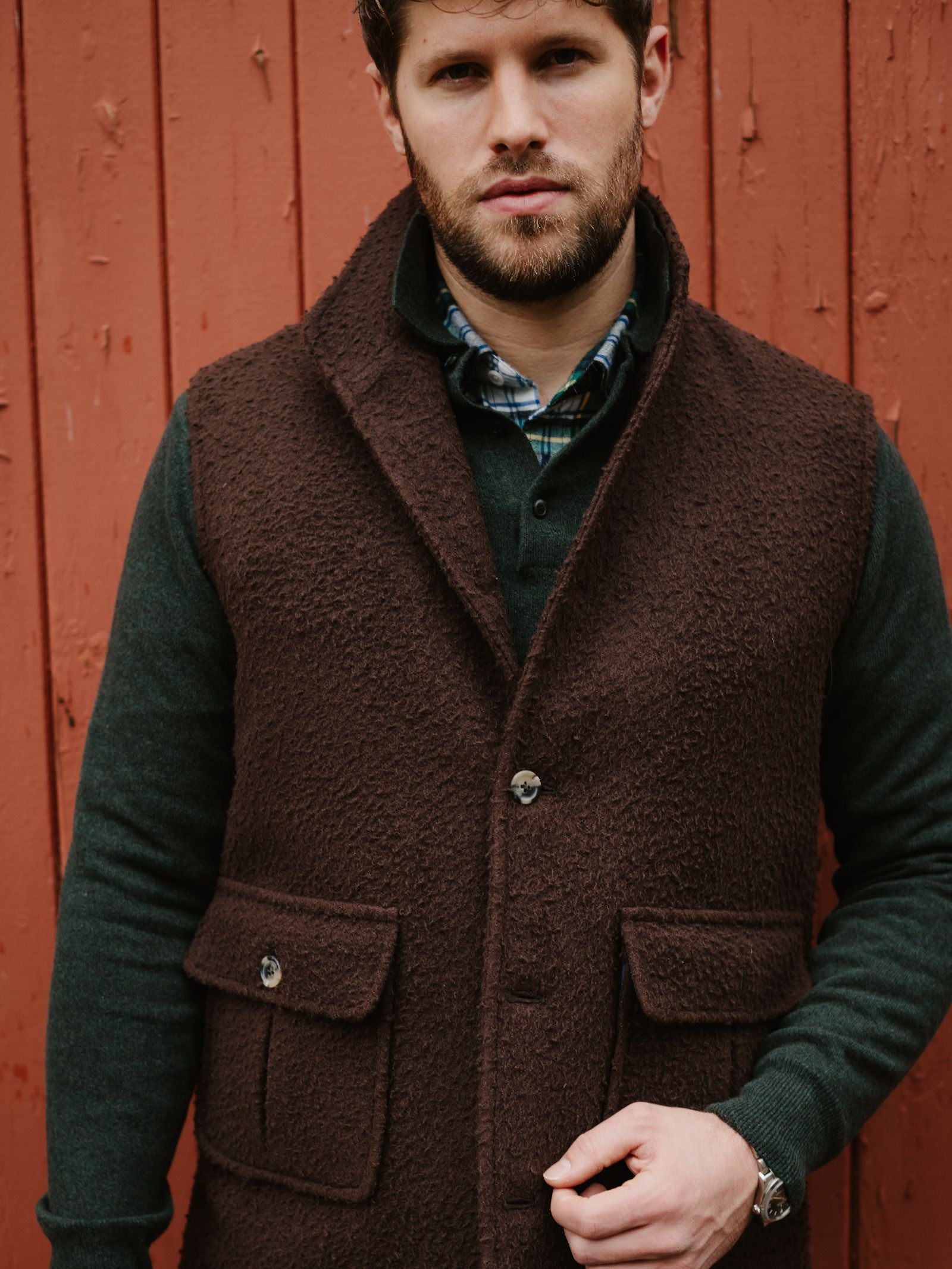 A man with short brown hair and a beard stands before a red wooden wall, wearing the Casentino Fleece Gilet by Campbell's of Beauly over a green long-sleeve shirt with a plaid shirt underneath.