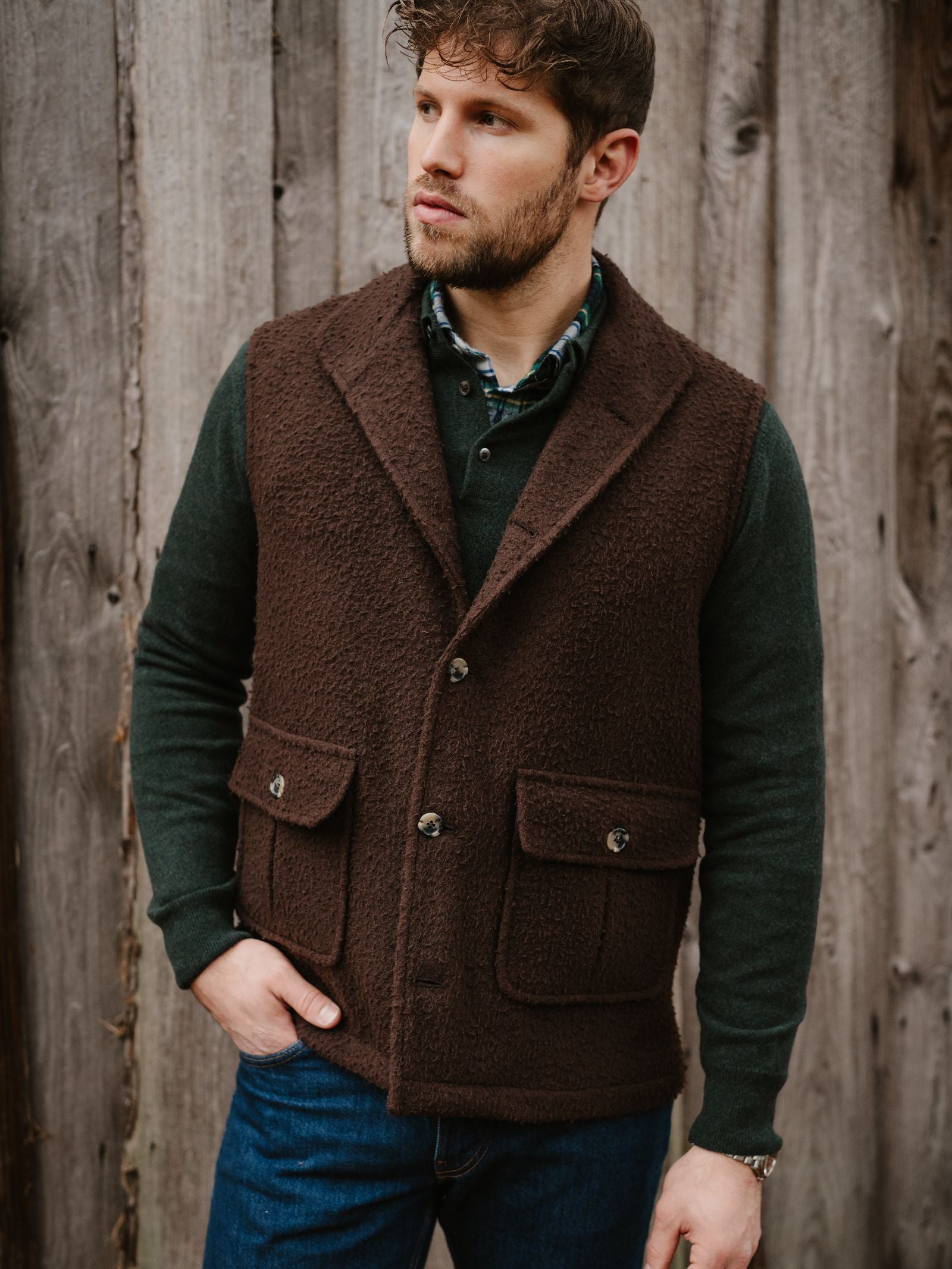 A man with short, curly brown hair and a beard stands before a wooden wall, wearing a green shirt, blue jeans, and the Campbell's of Beauly Casentino Fleece Gilet with pockets and buttons, gazing off to the side.