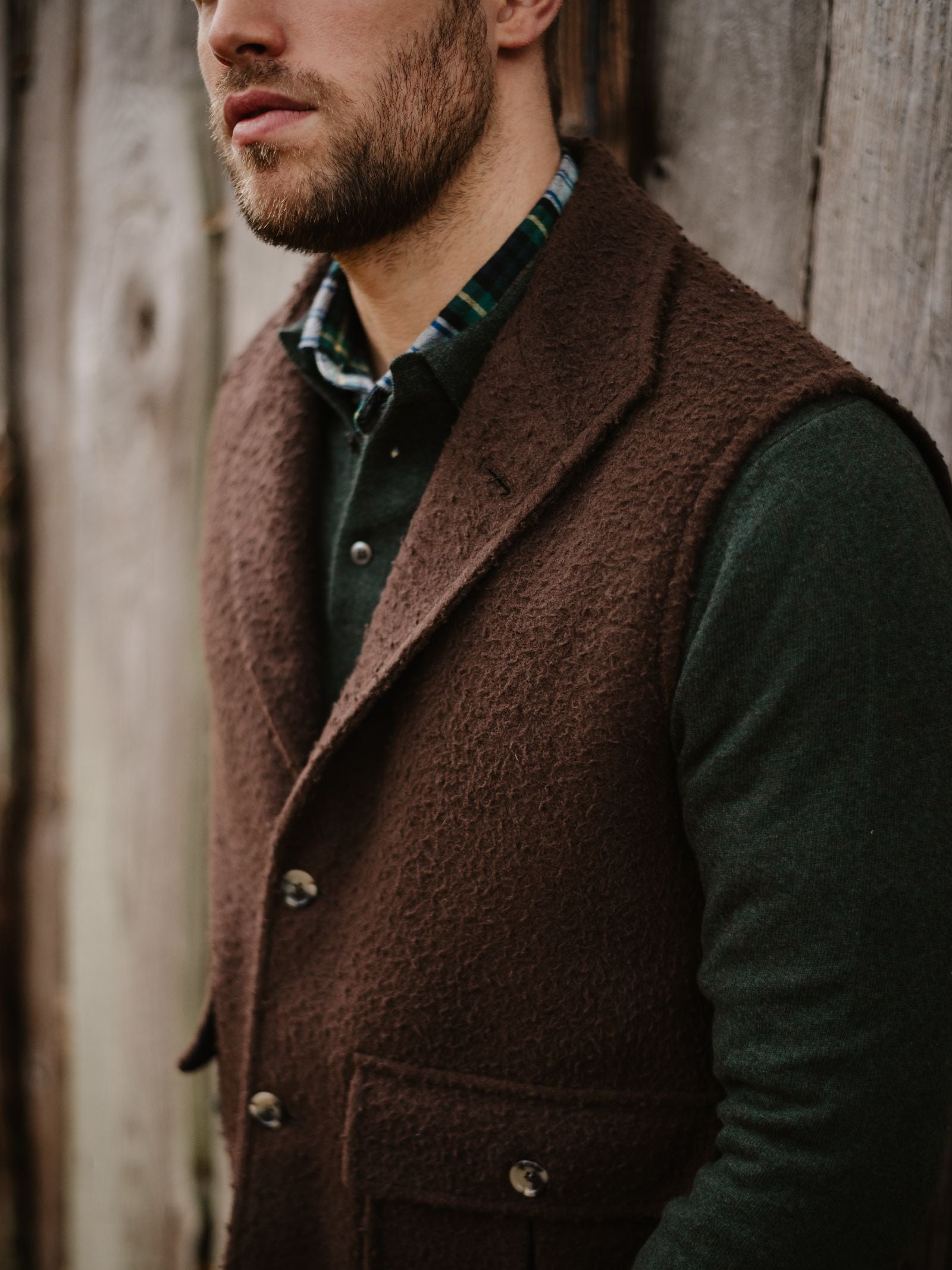 A bearded man in a dark green shirt, green plaid collar, and Campbell’s of Beauly Casentino Fleece Gilet stands before a rustic wooden wall. His face is partially out of frame.