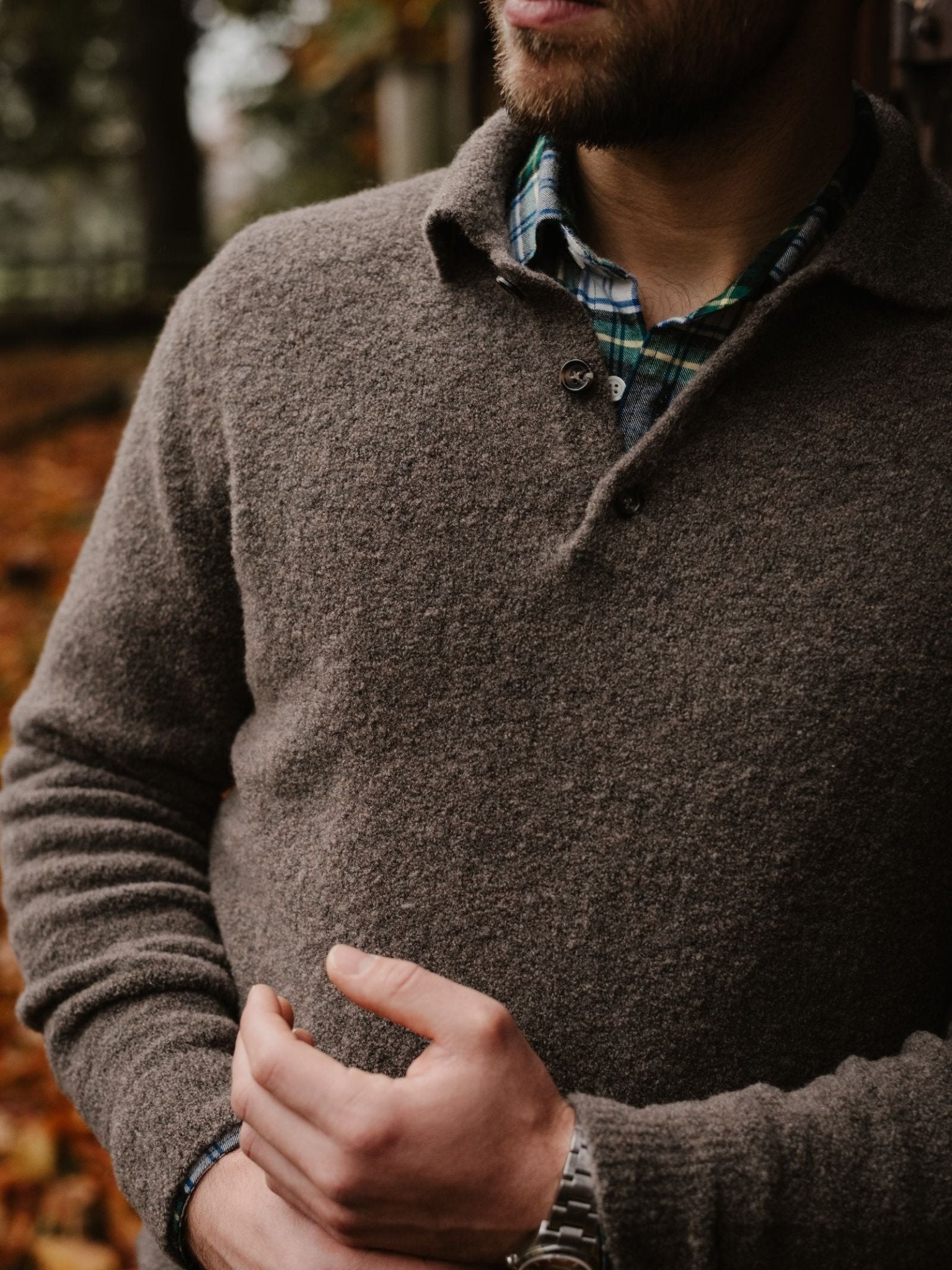 A bearded man stands outdoors among autumn leaves, wearing a brown Campbell's of Beauly Bouclé Polo Sweater over a green and blue plaid shirt, with his hand resting near his waist.