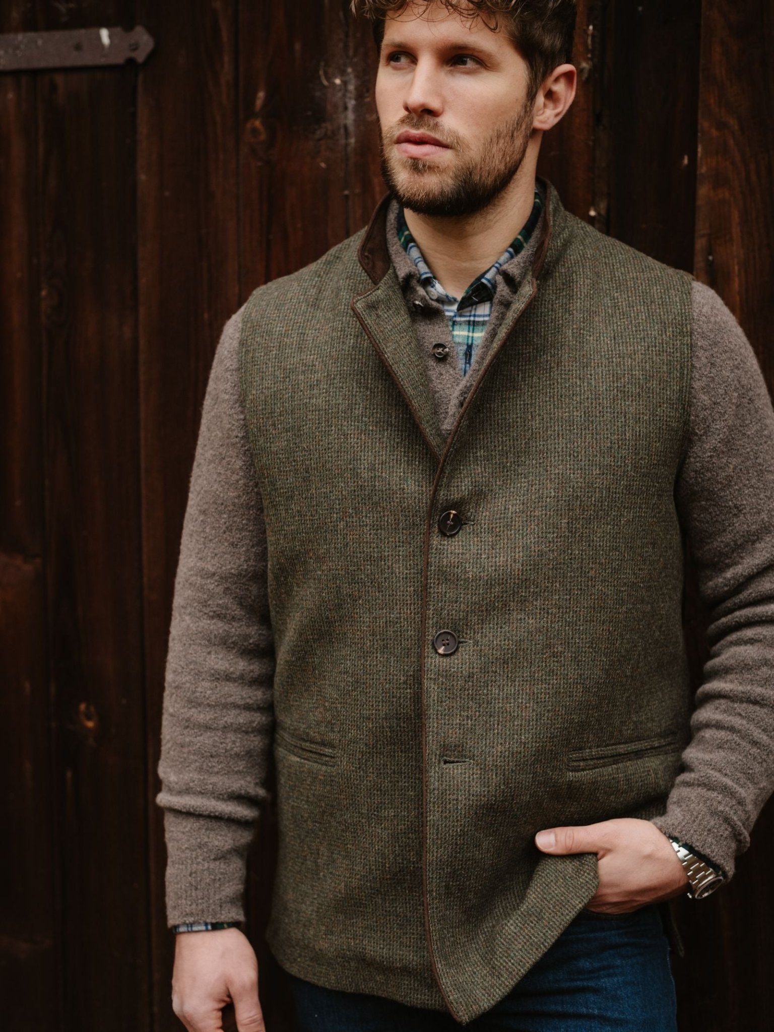 A man wears the Campbell's of Beauly Tweed Nehru Gilet over a gray sweater and plaid shirt, standing before a dark wooden wall—an elegant layering piece for refined style.