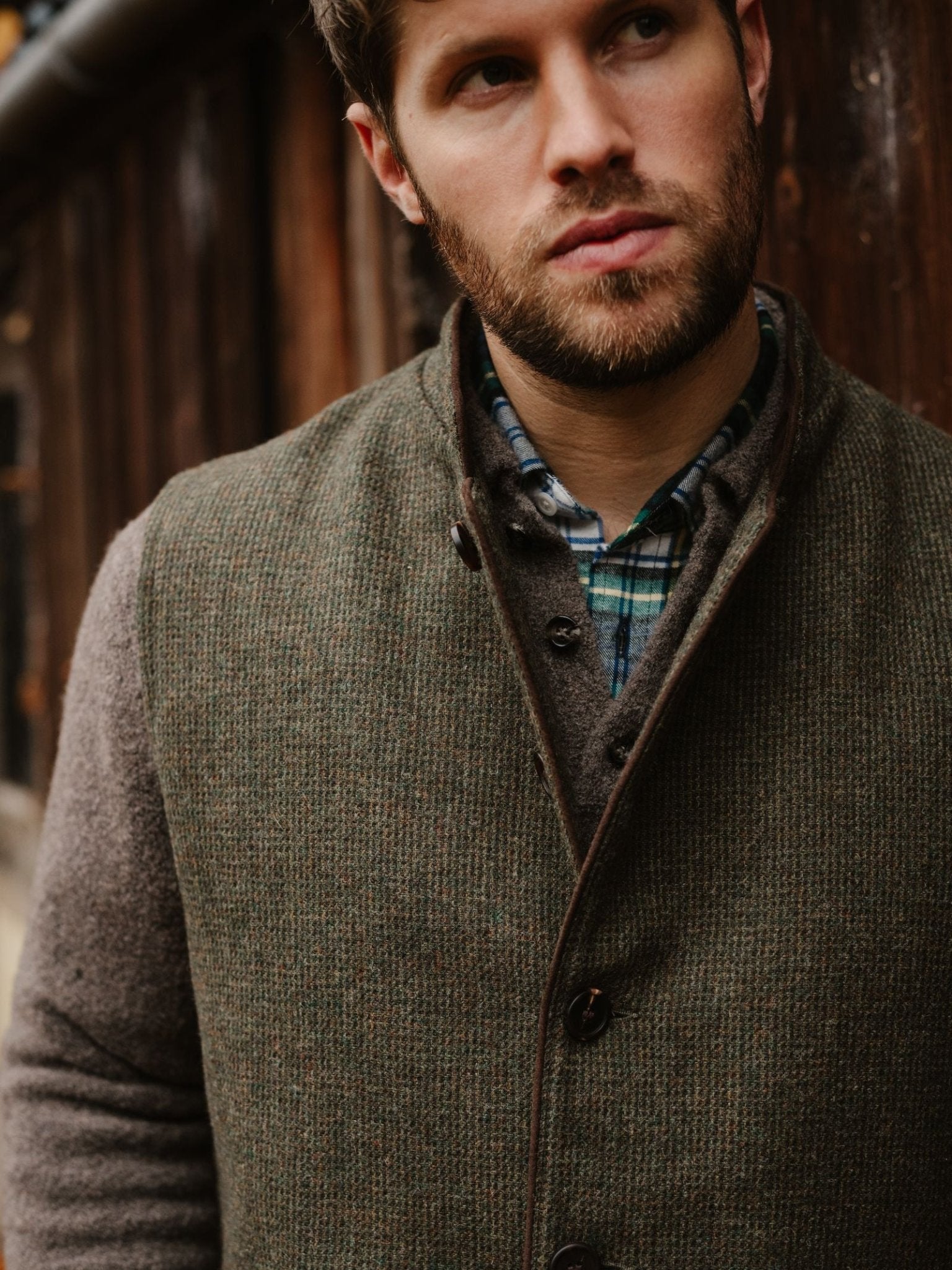 A bearded man in a plaid shirt and Campbell's of Beauly Tweed Nehru Gilet stands by a wooden wall, looking thoughtfully to the side—a perfect example of classic layering style.