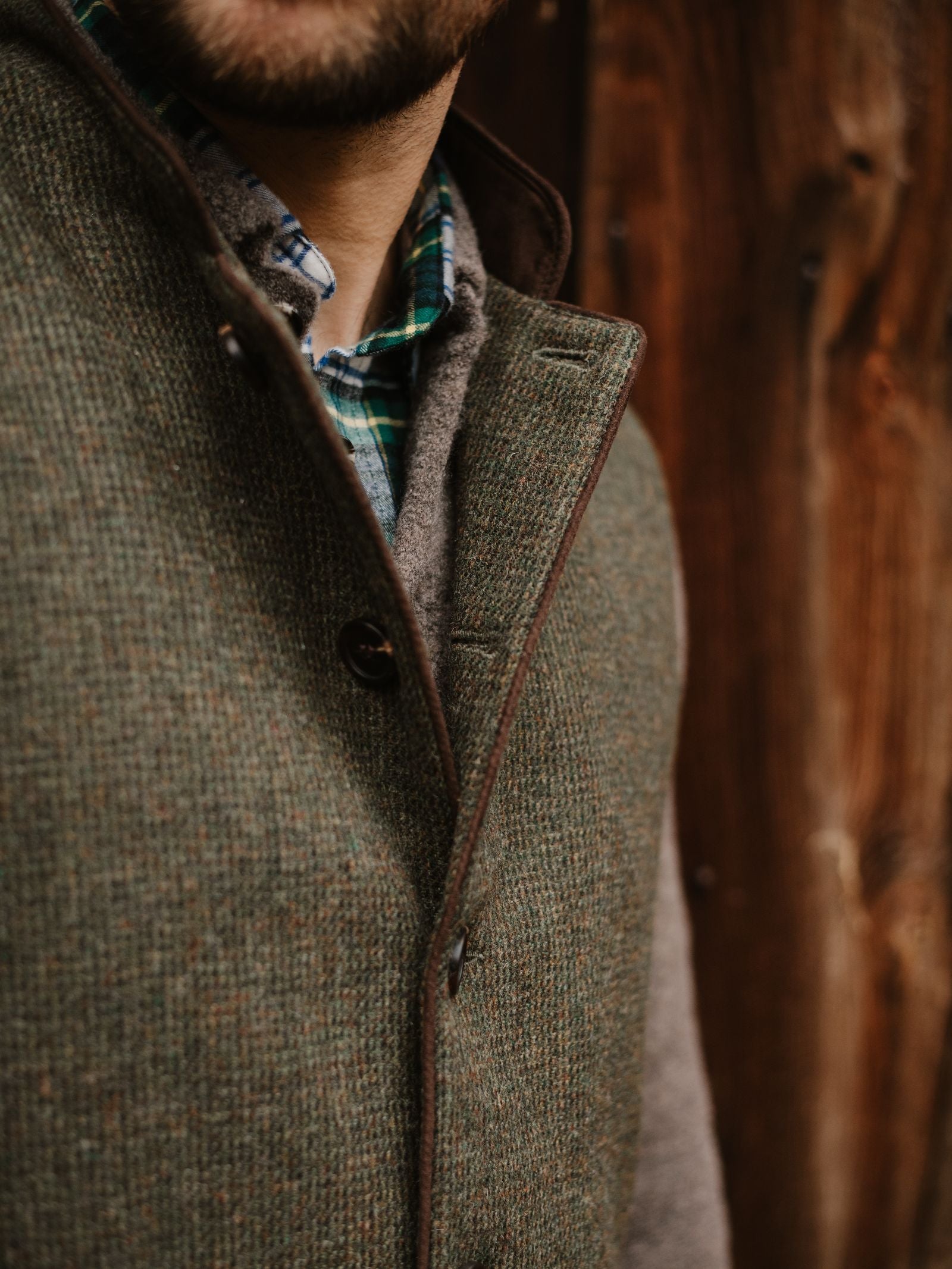 A close-up of a man wearing the Campbell's of Beauly Tweed Nehru Gilet over a blue and green plaid shirt, standing in front of a rustic wooden wall, with only his lower face and upper torso visible.