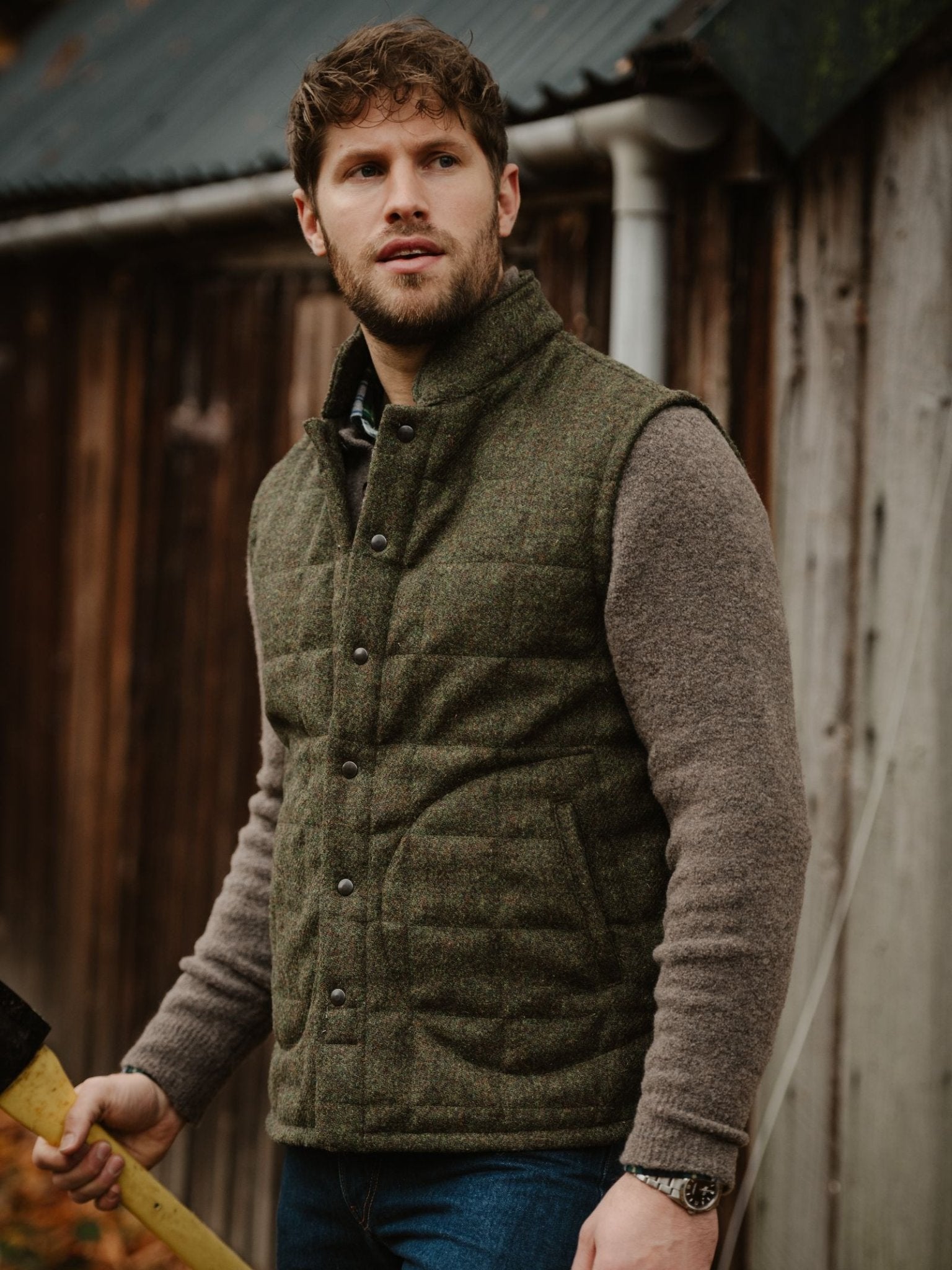 A bearded man stands outside a weathered wooden building, holding an axe and wearing the Campbell's of Beauly Harris Tweed Quilted Gilet over a brown sweater and blue jeans, looking off to the side.