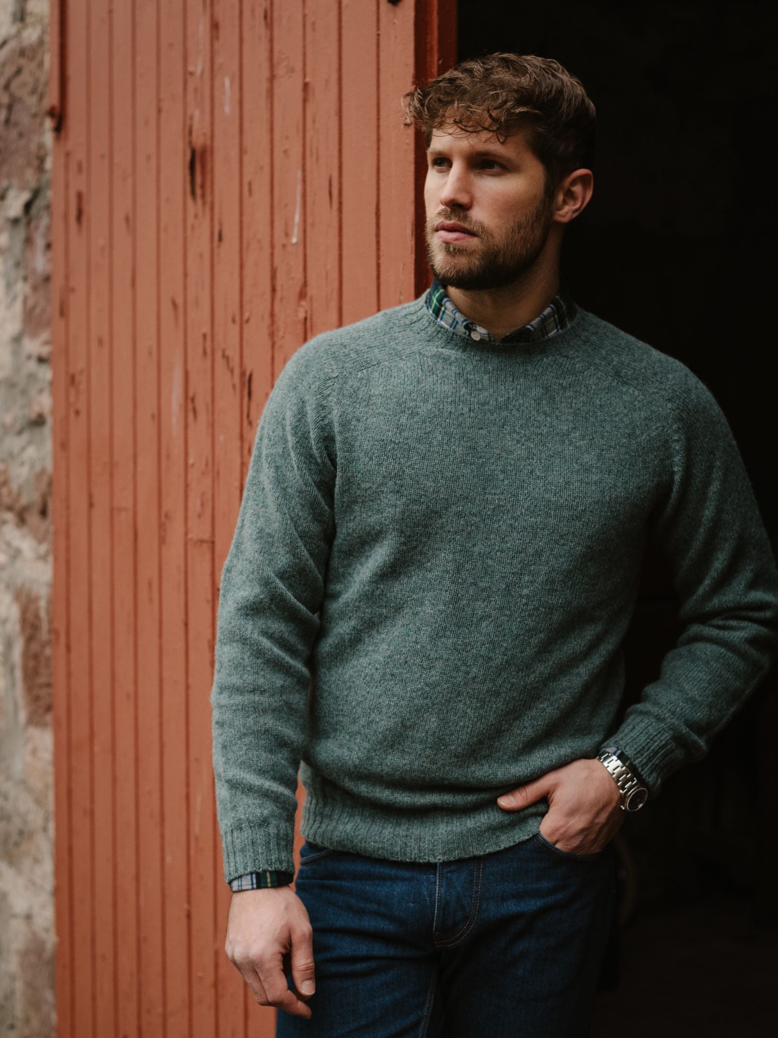 A man with short curly hair and a beard stands by a rustic red door, wearing a Campbell's of Beauly Shetland Jumper in green over a plaid shirt and blue jeans, with one hand in his pocket and a wristwatch showing.