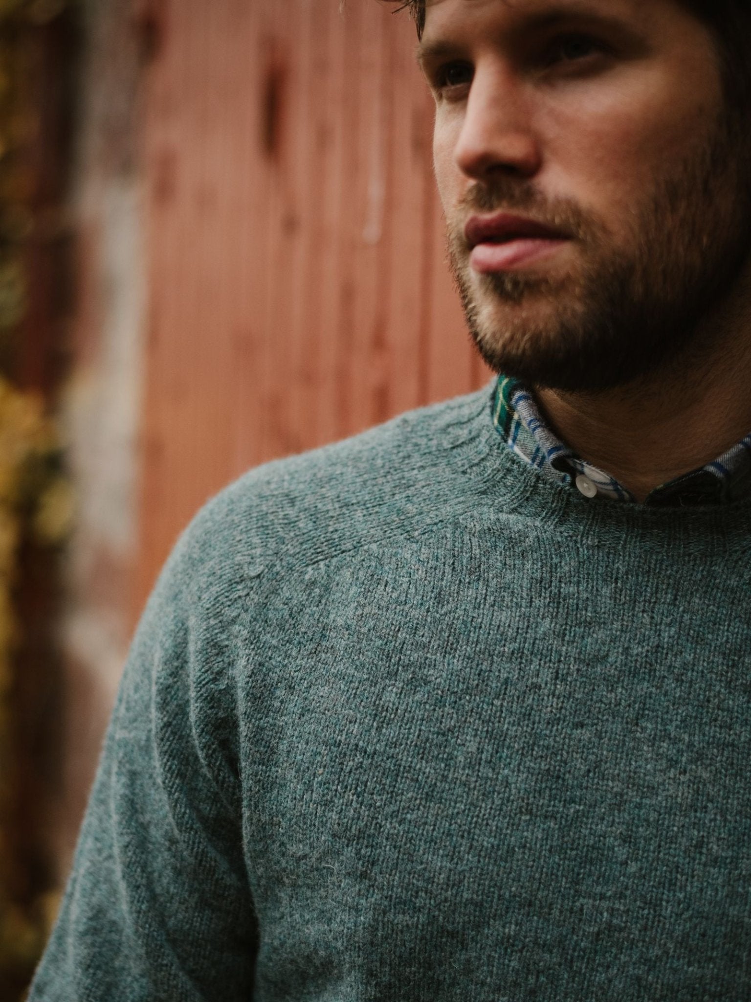 A bearded man wearing a green Campbell's of Beauly Shetland Jumper over a plaid shirt stands in front of a wooden wall, gazing to the side. Only his upper body and part of his face are visible.