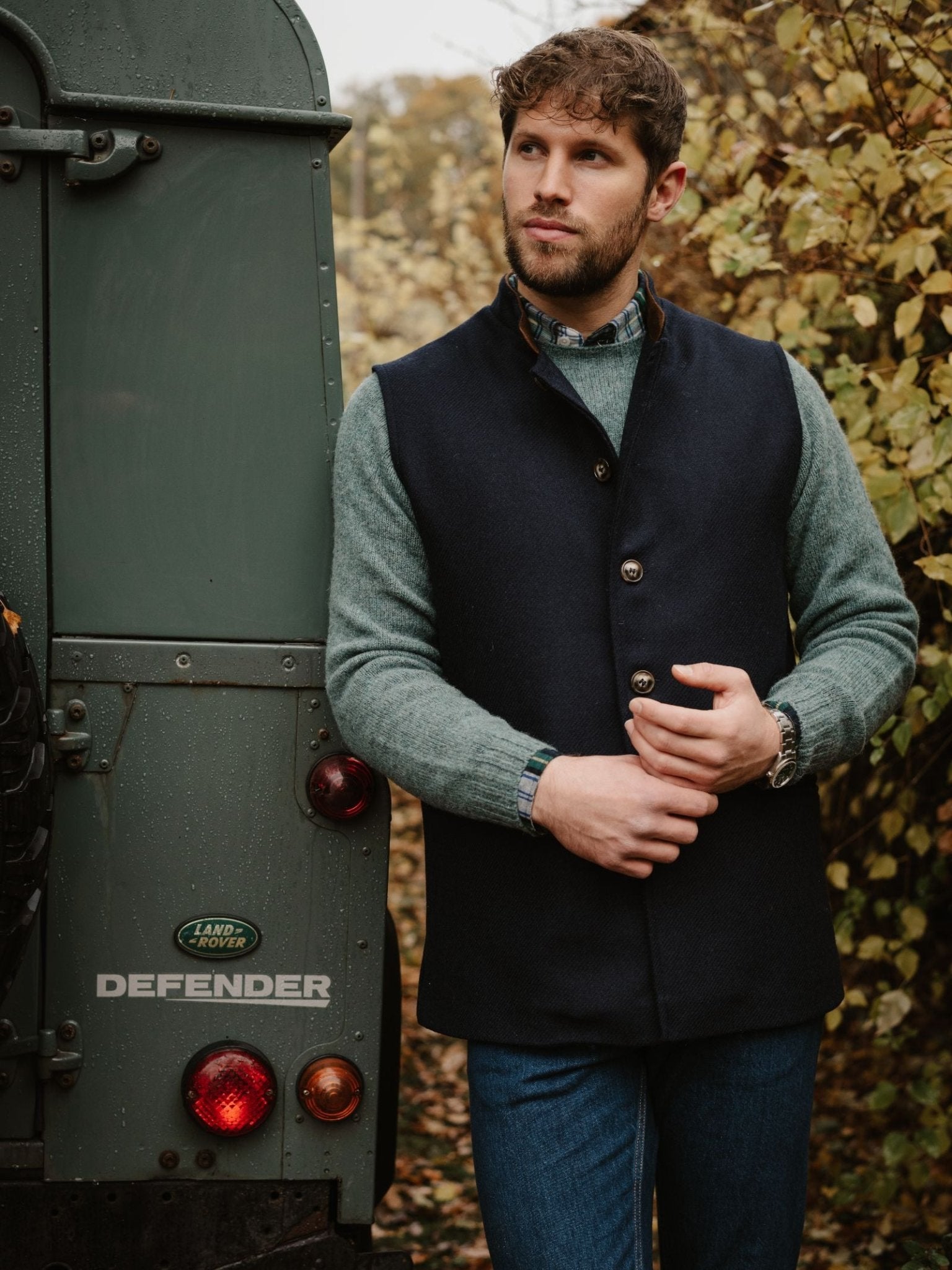 A bearded man stands outdoors by a green Land Rover Defender, wearing the Campbell's of Beauly Tweed Nehru Gilet layered over a green sweater and blue jeans, with autumn leaves in the background.