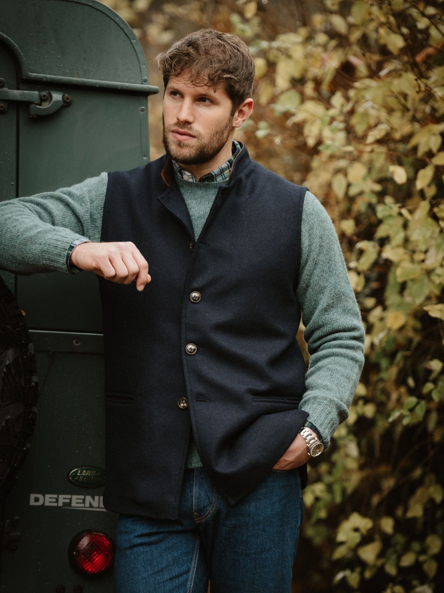 A man with short brown hair and a beard stands outdoors by a green vehicle, wearing a Campbell's of Beauly Tweed Nehru Gilet over a green sweater, blue jeans, and a watch, with autumn leaves in the background.