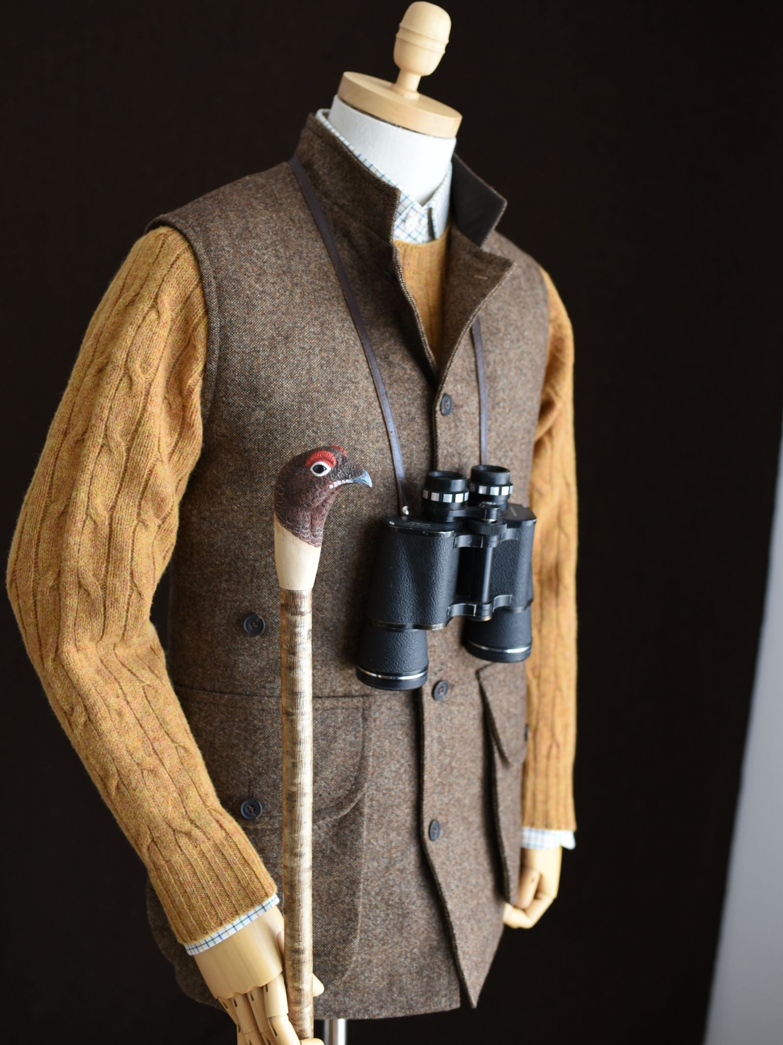 A mannequin wears a Campbell's of Beauly Auld Stock Field Nehru Gilet over a mustard cable-knit sweater, holds a bird-handle walking stick, and has black binoculars hanging from its neck.