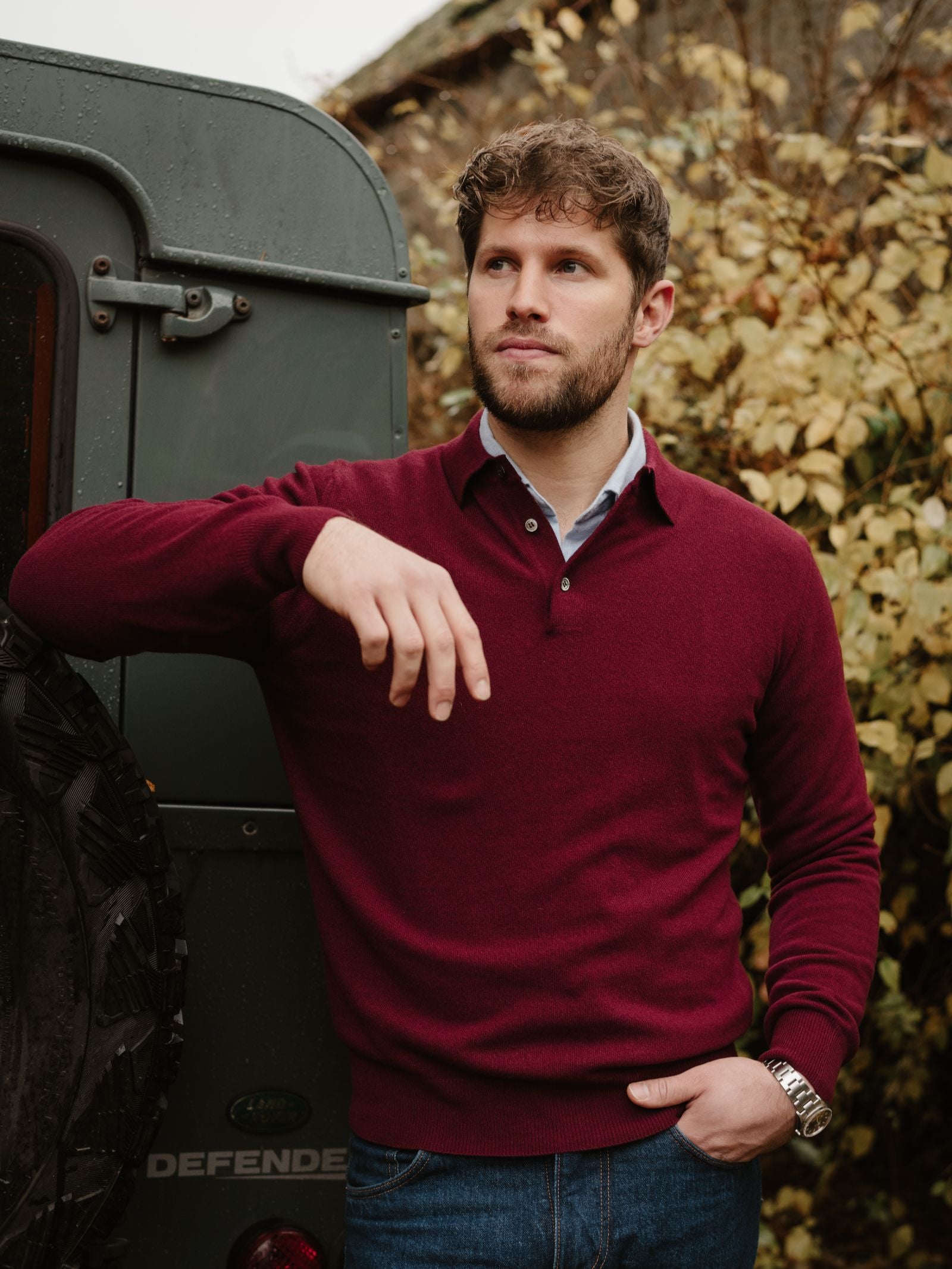 A man with short curly hair and a beard, wearing Campbell's of Beauly's Merino & Cashmere Shirt Jumper in maroon and blue jeans, leans against a dark green Land Rover Defender amid vibrant autumn foliage.