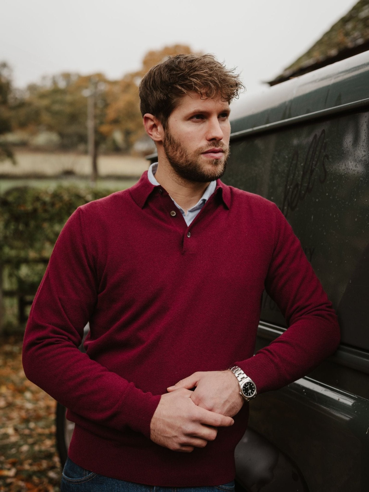 A man with short brown hair and a beard wears the Campbell’s of Beauly Merino & Cashmere Shirt Jumper—maroon over a blue shirt—standing outdoors by a green vehicle amid autumn trees and fallen leaves.